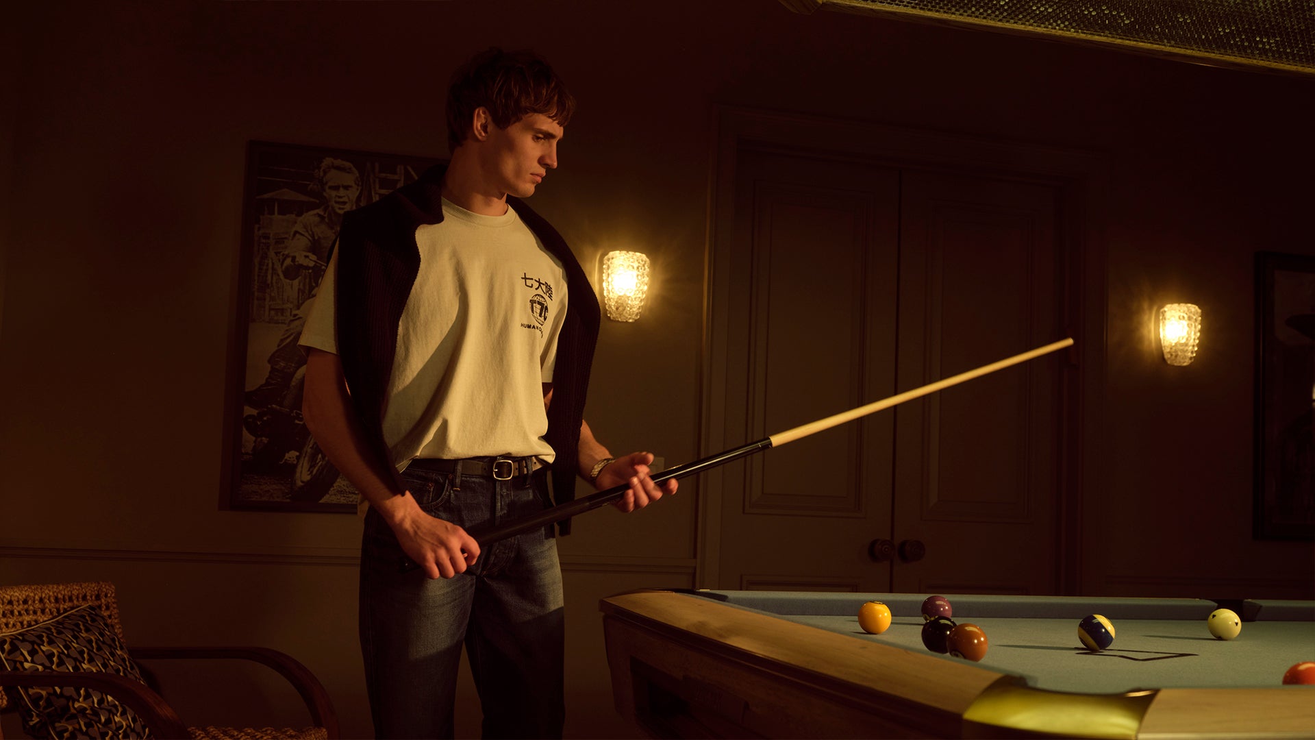 A male model wearing a t-shirt and jeans playing pool in a dark room