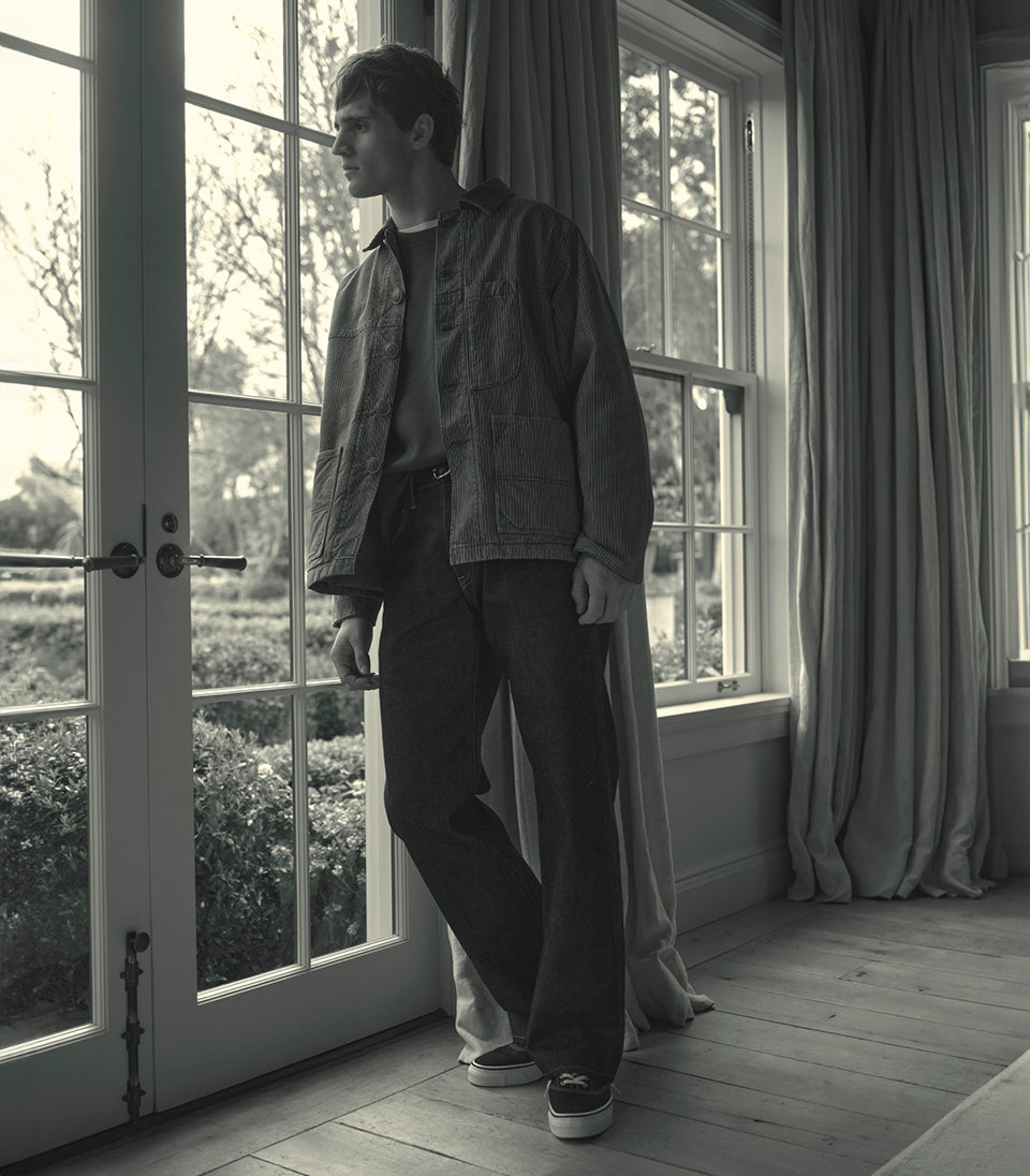 A male model wearing a jacket and relaxed denim jeans staring out a window