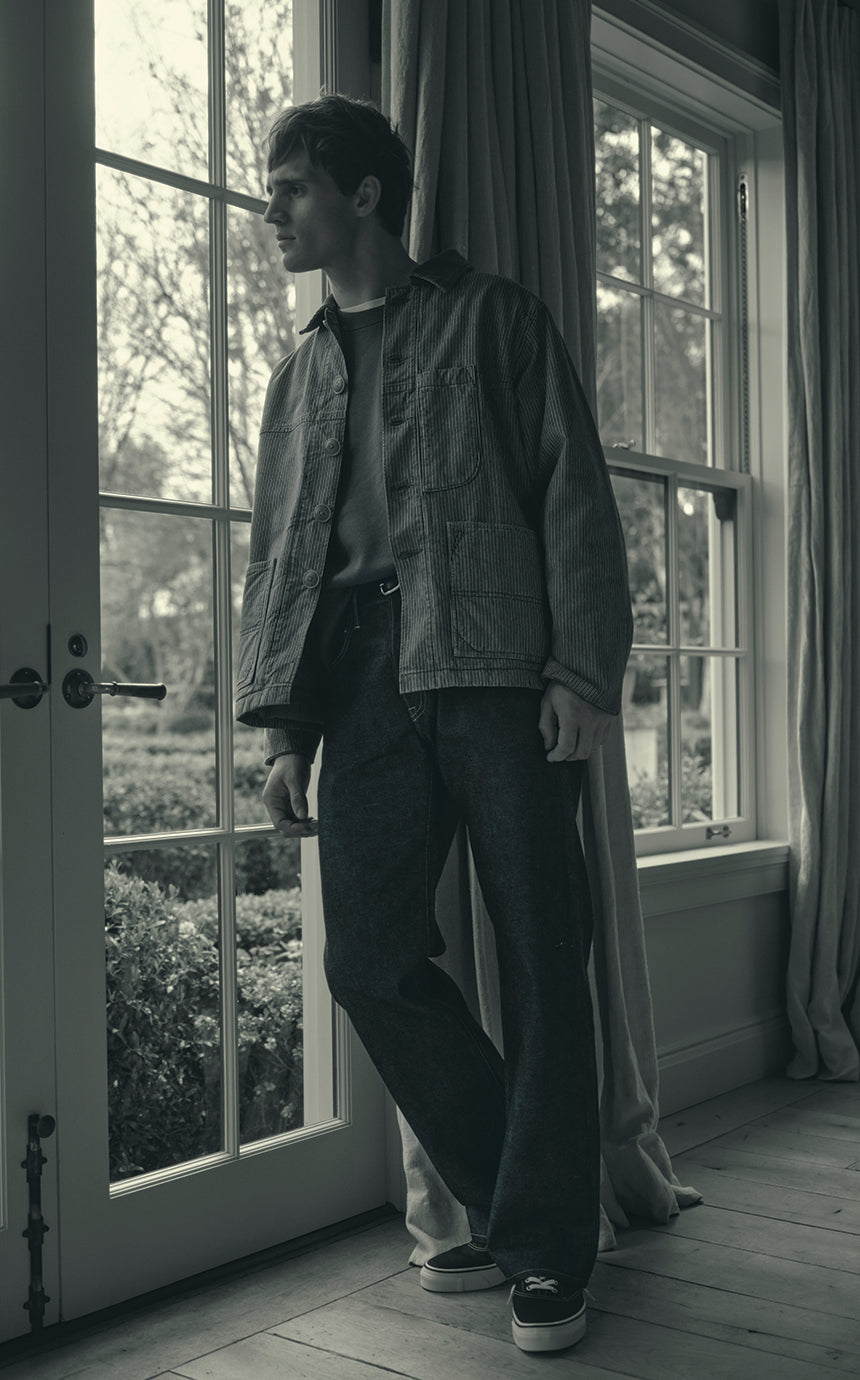 A male model wearing a jacket and relaxed denim jeans staring out a window