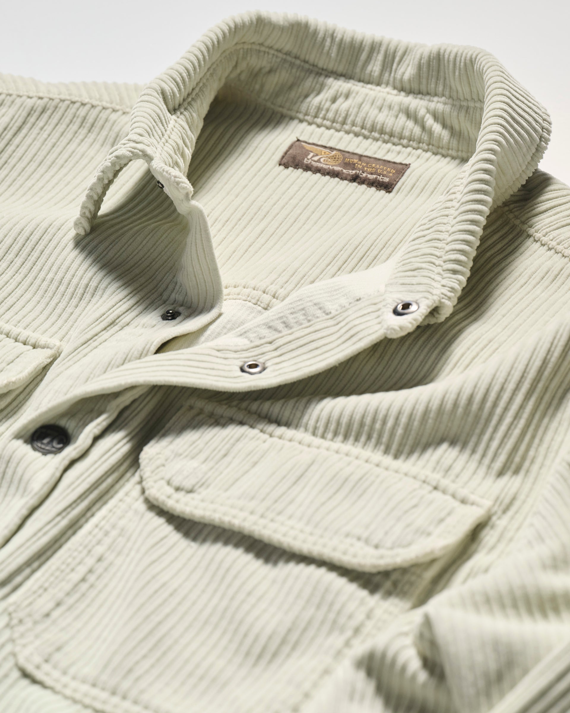 A close-up of The Aiden Overshirt in Cream by T7C, featuring a collar, two front pockets, metal snap buttons, a soft corduroy texture, and a visible inside collar brand label that reflects its made-in-LA craftsmanship.