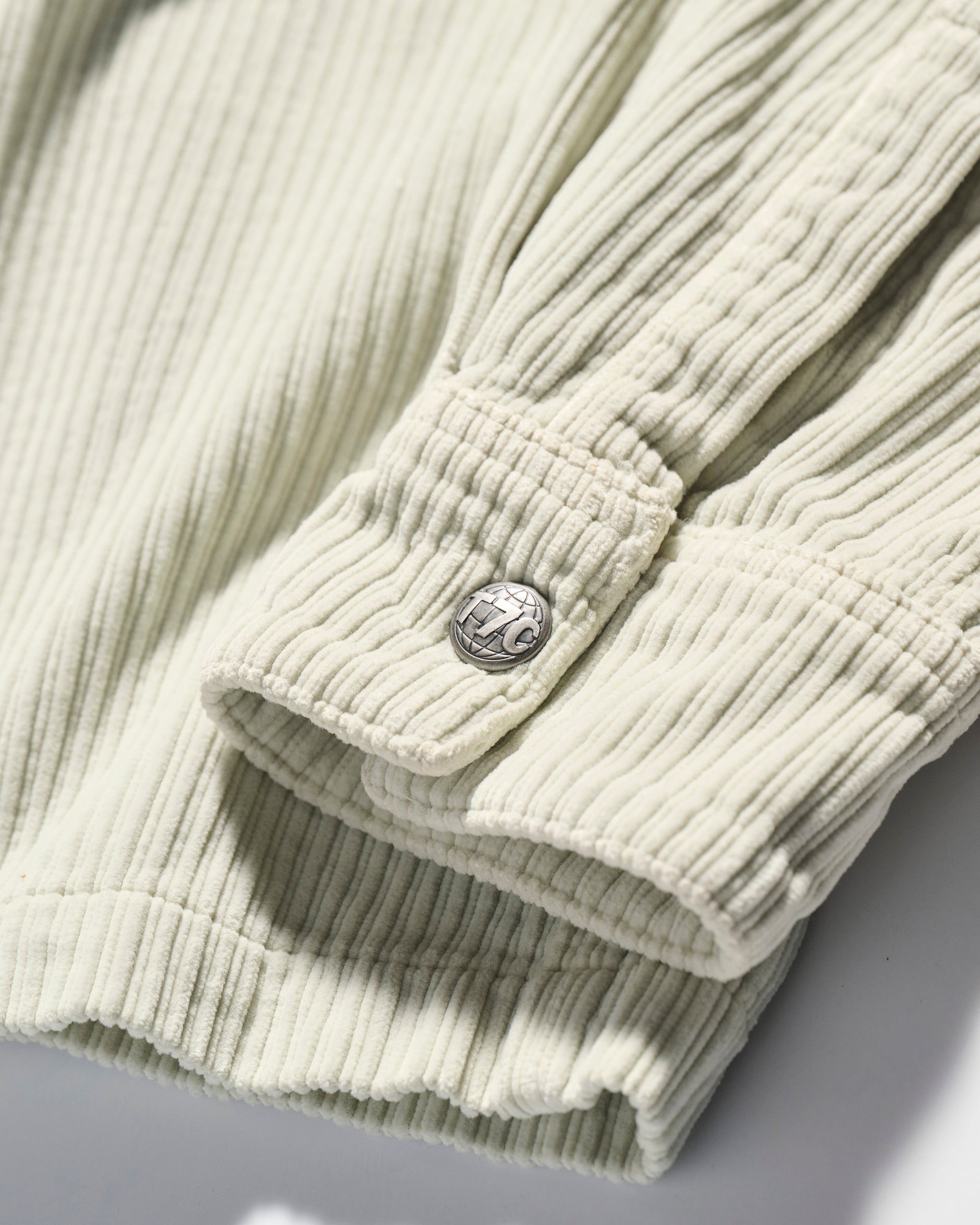 Close-up of The Aiden Overshirt in Cream by T7C, showcasing its ribbed Japanese corduroy sleeve and a metal button embossed with a mountain and wave. The soft, textured fabric makes it ideal for a relaxed look, with button detail sharply in focus.