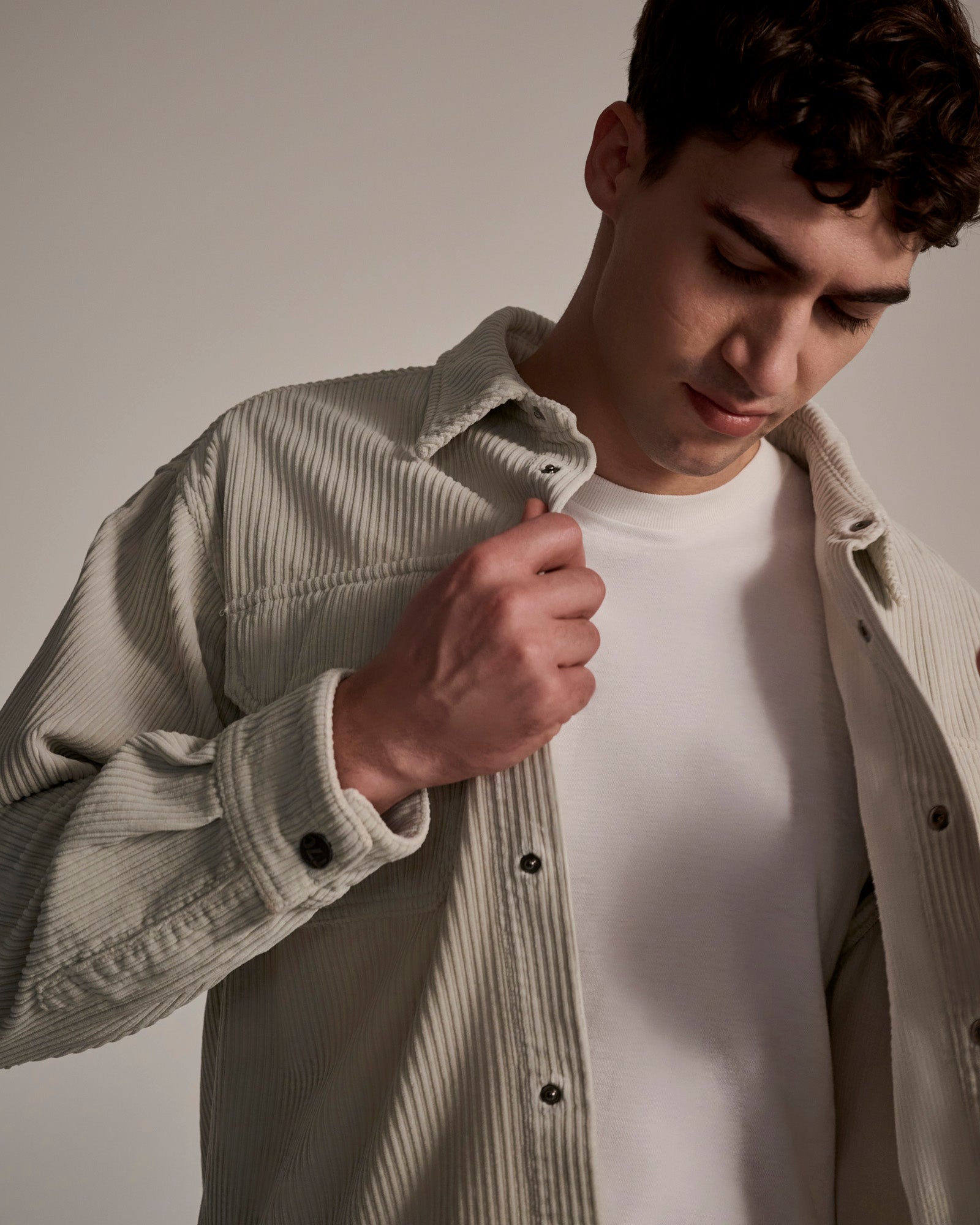 A young man wearing a white t-shirt and The Aiden Overshirt in Cream by T7C holds the collar with one hand, looking down against a neutral background.