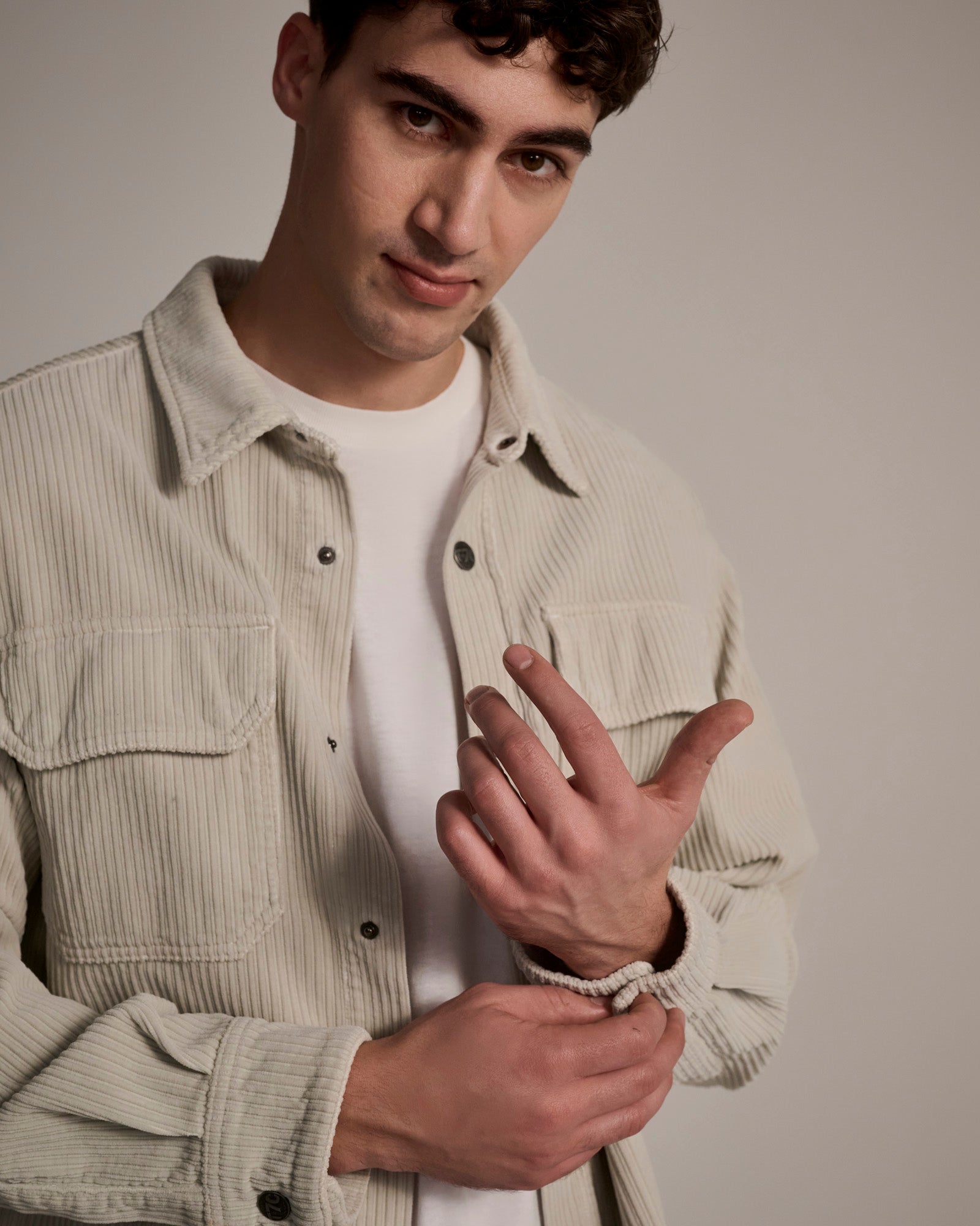 A young man with short dark hair wears The Aiden Overshirt in Cream by T7C over a white t-shirt, holding his left wrist with his right hand and smiling slightly at the camera against a plain background.