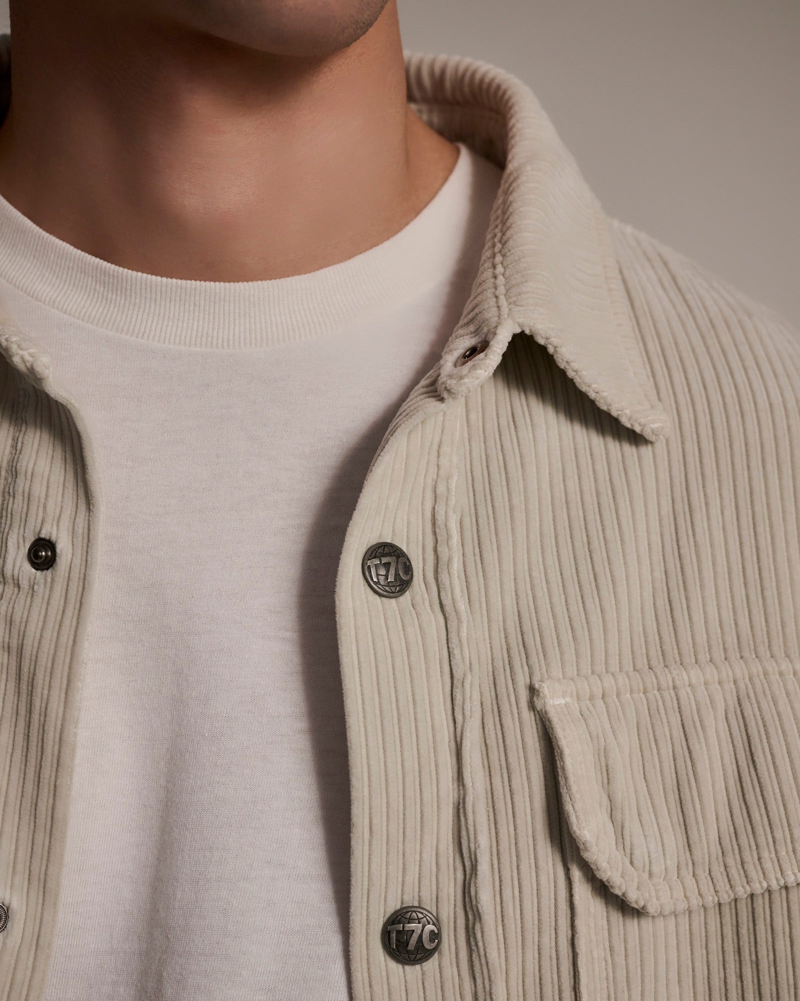 Close-up of a person wearing T7Cs The Aiden Overshirt in Cream, featuring Japanese corduroy and metal buttons, layered over a white crew-neck t-shirt; only the lower face, neck, and upper torso are shown.