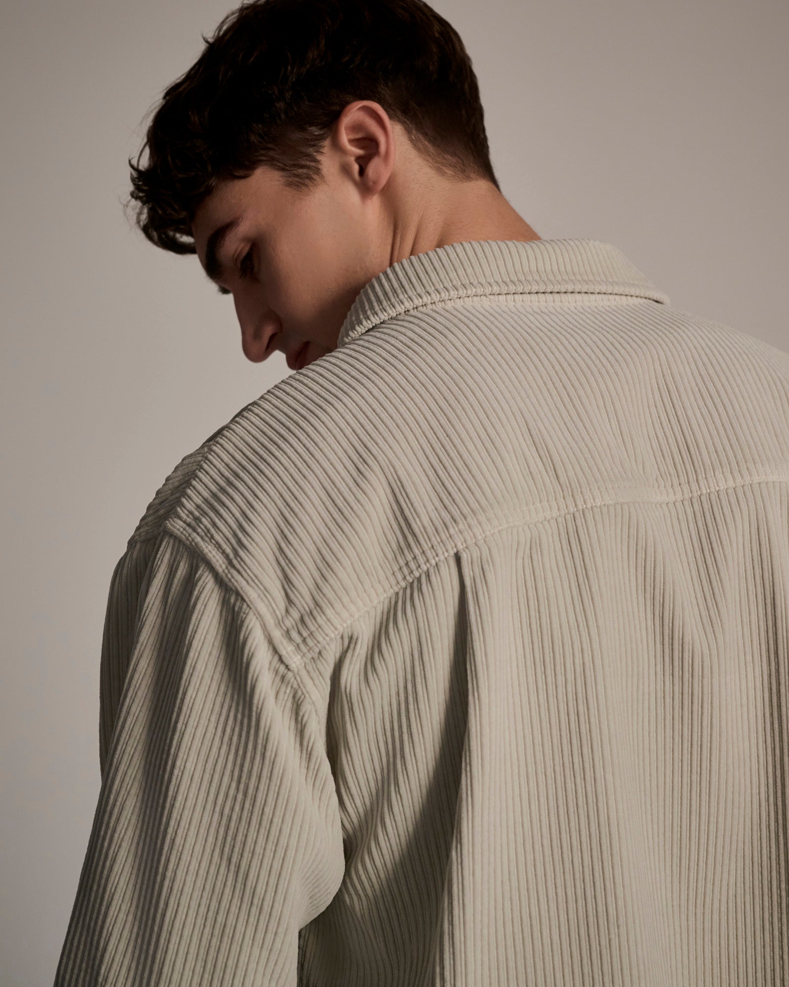 A young man with short brown hair is shown from behind, wearing The Aiden Overshirt in Cream by T7C. He slightly turns his head down and to the side, set against a soft, neutral background.
