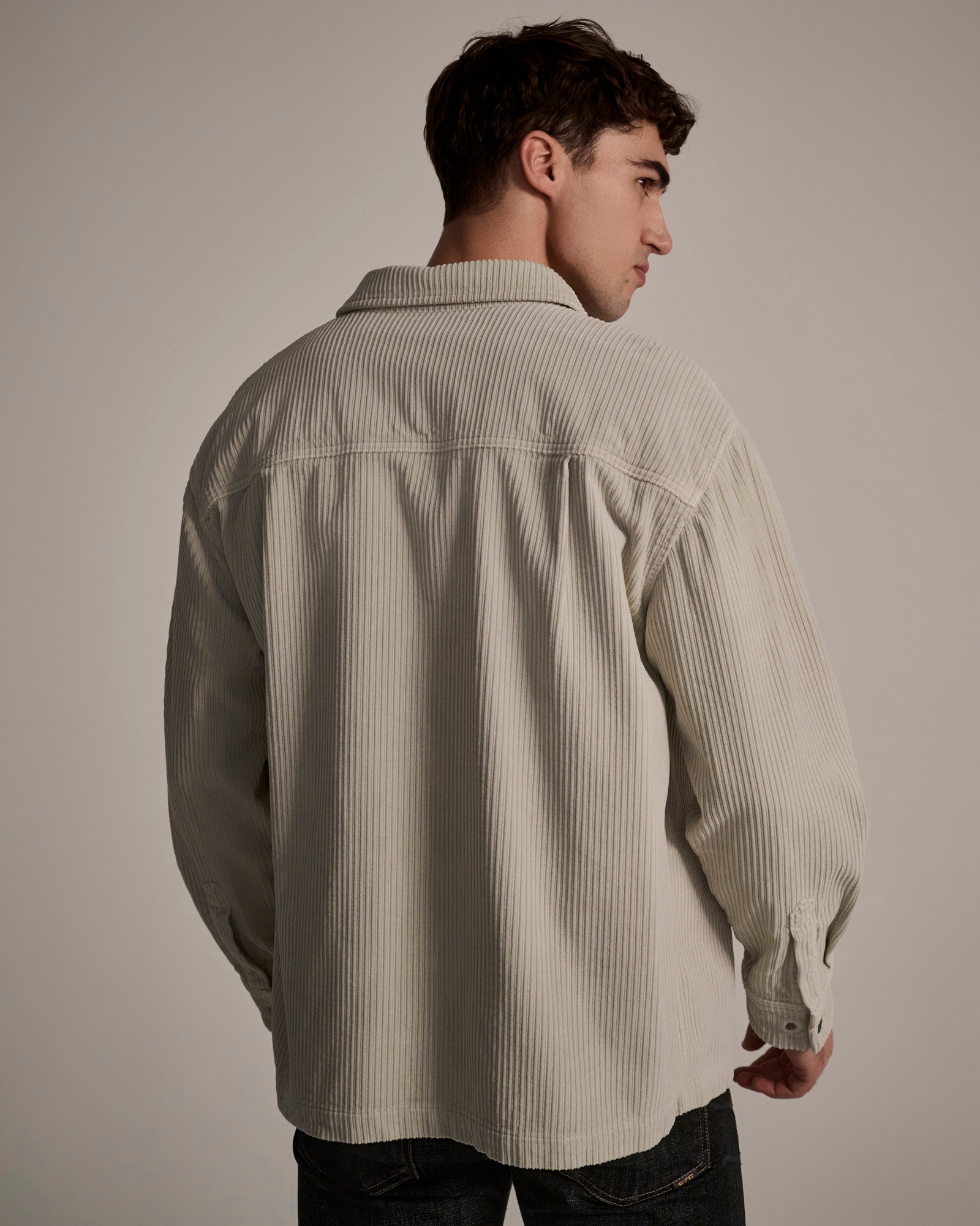 A young man with short brown hair is seen from behind, wearing T7Cs The Aiden Overshirt in Cream paired with dark jeans, standing against a plain beige background.