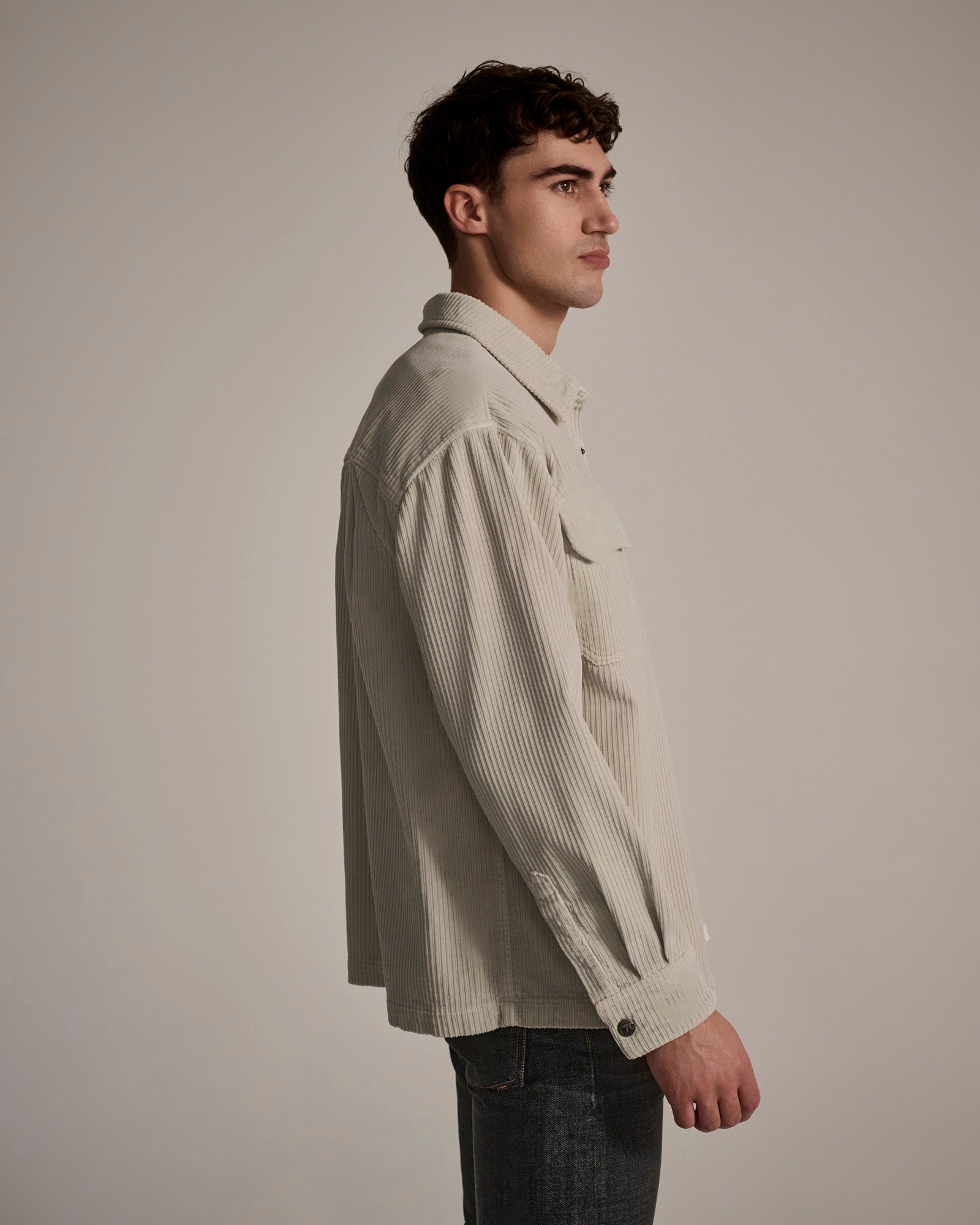A man with short brown hair models the T7C Aiden Overshirt in Cream, paired with dark jeans, standing in profile against a plain, neutral background.
