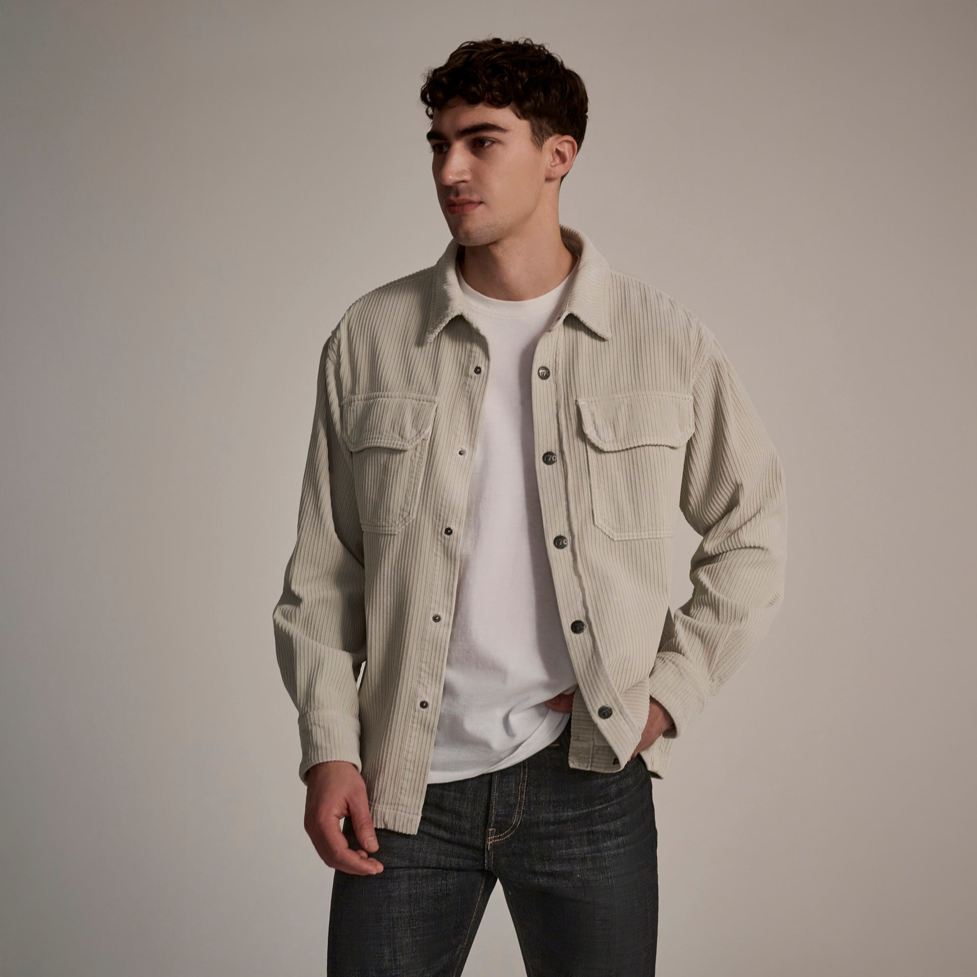 A young man stands against a plain background, wearing The Aiden Overshirt in Cream by T7C over a white t-shirt and dark jeans. He looks slightly to his left with one hand in his pocket.