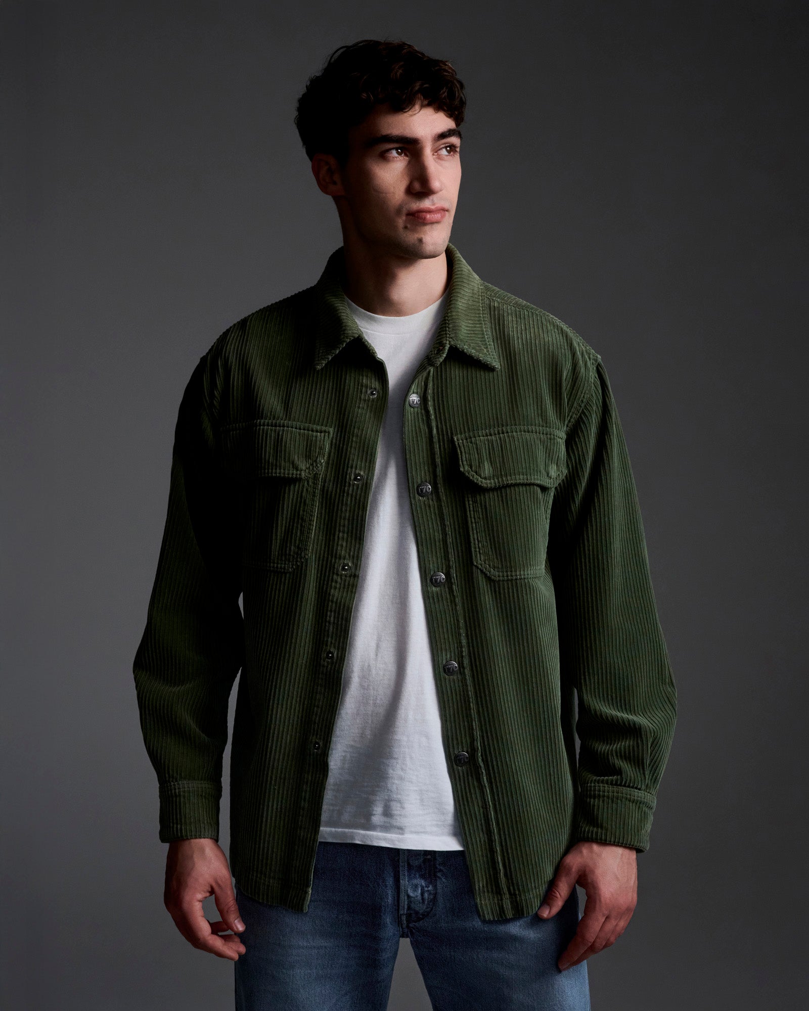 A young man with short dark hair wears T7C’s The Aiden Overshirt in Green, made in LA, over a white t-shirt and blue jeans as he stands against a plain, dark background, looking slightly to his right.