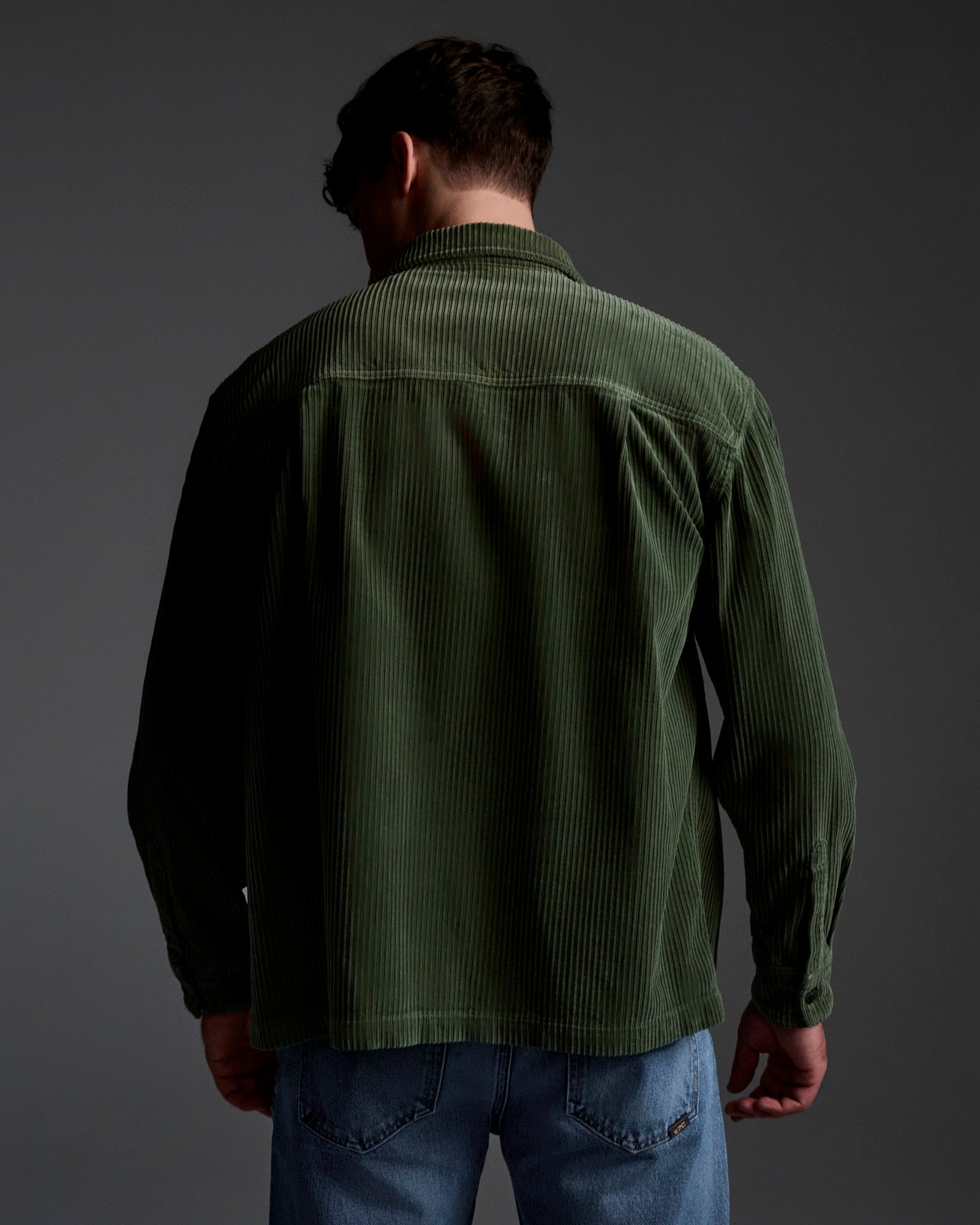 A person stands with their back to the camera in blue jeans and T7C’s The Aiden Overshirt in Green, set against a dark gray backdrop. Soft lighting accentuates the overshirt’s textured fabric.