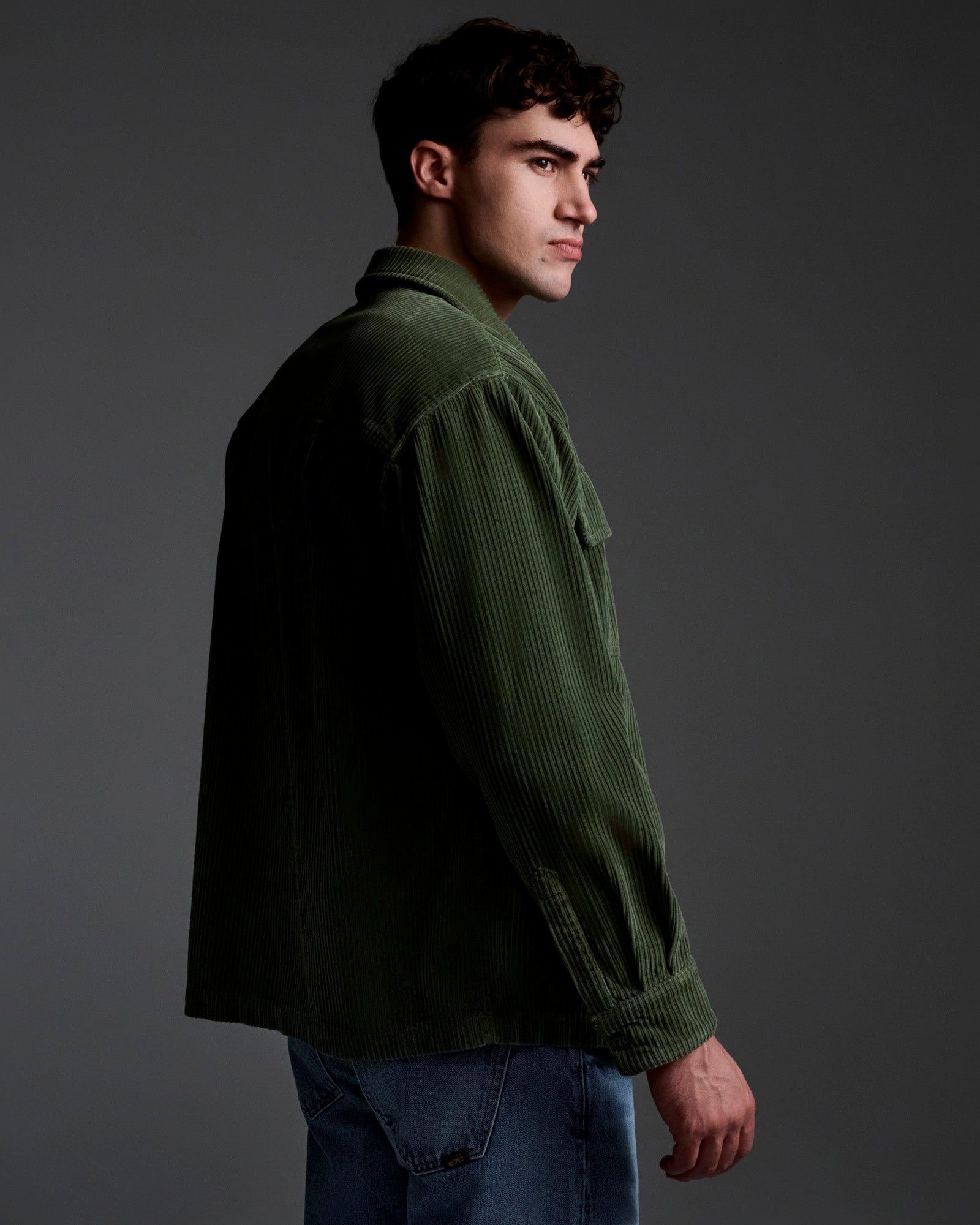 A young man stands in profile against a dark background, wearing T7C’s The Aiden Overshirt in Green and blue jeans, looking ahead with a neutral expression.