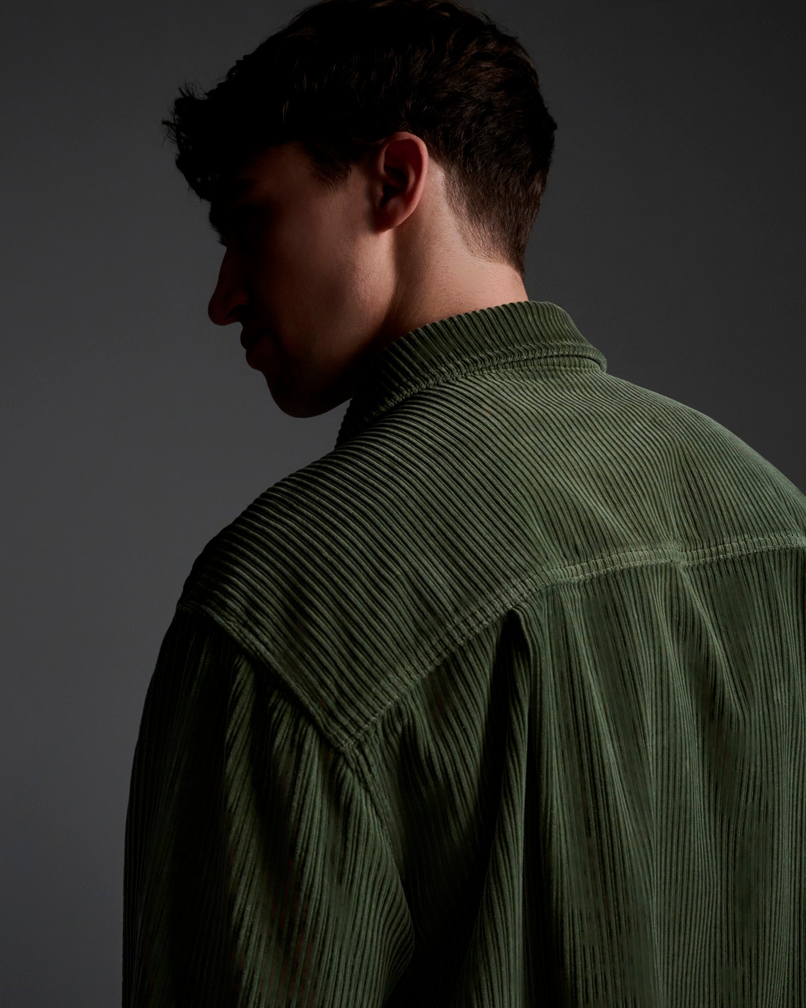 A man is seen from behind and slightly in profile, wearing T7Cs The Aiden Overshirt in Green. Dim lighting and a plain gray background create a moody atmosphere.
