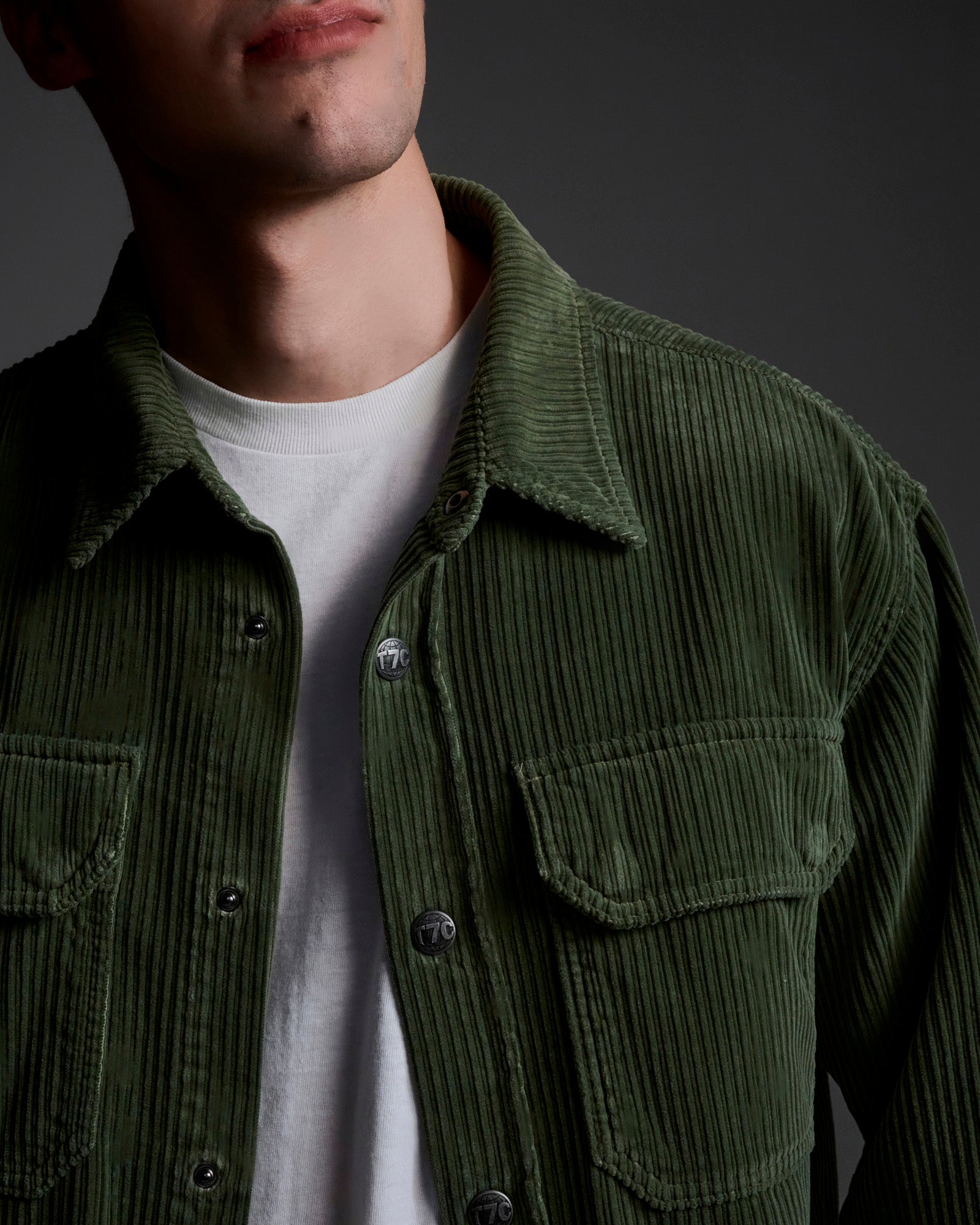 A person wears a white t-shirt and the T7C Aiden Overshirt in Green, featuring chest pockets, shown from shoulders to just above the mouth against a dark background.