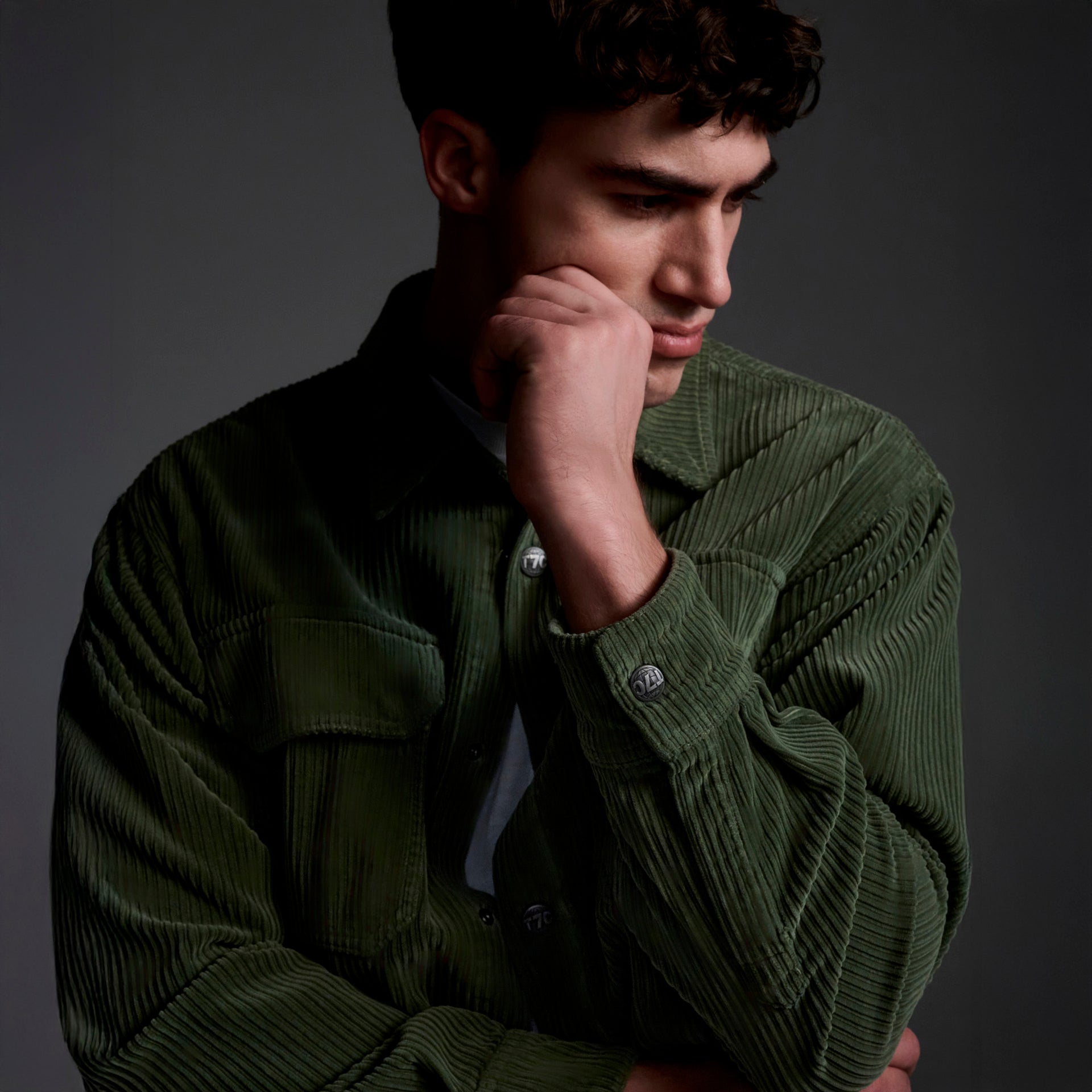 Wearing The Aiden Overshirt in Green by T7C, made in LA, a person stands against a dark background, gazing down thoughtfully with their hand on their chin.