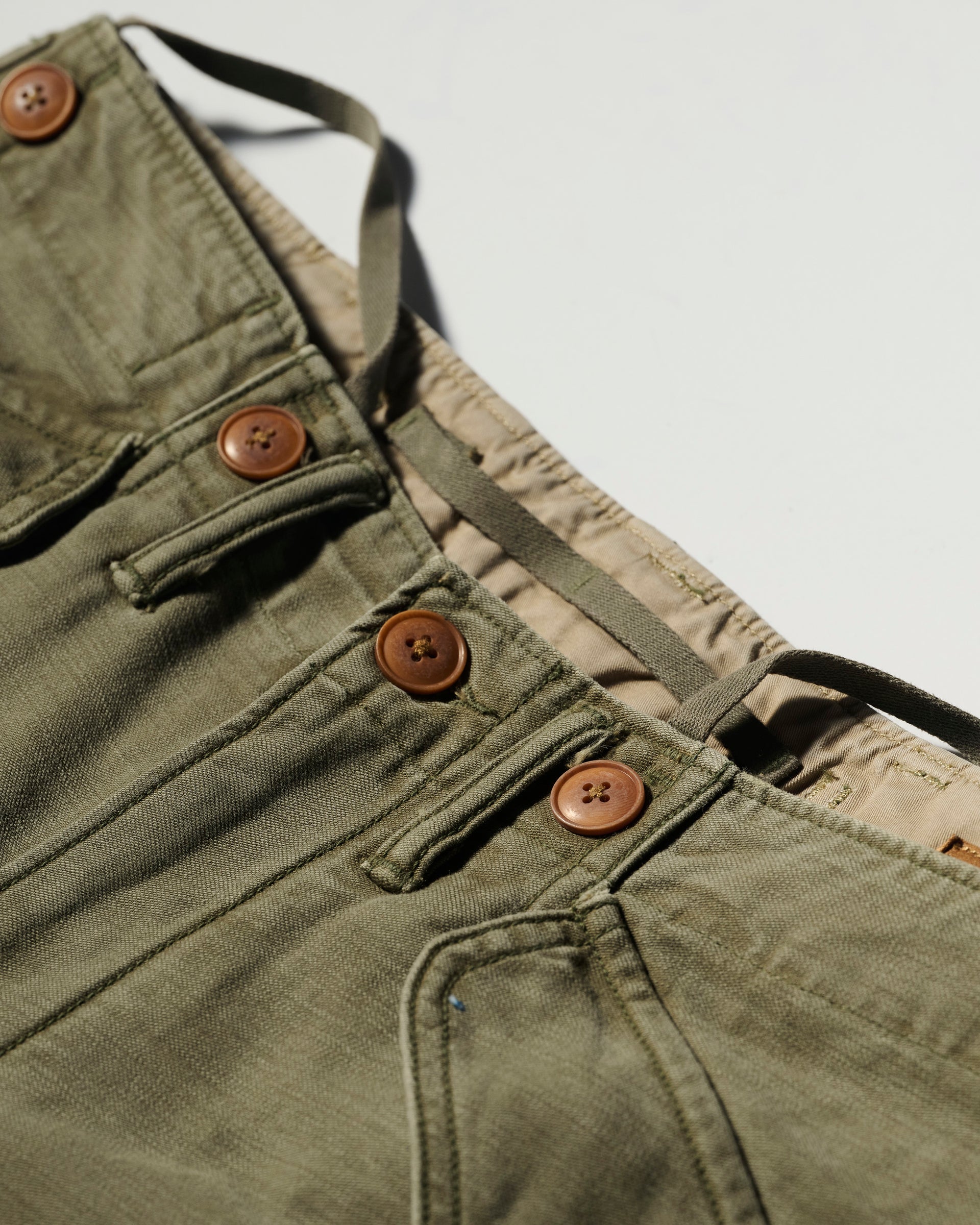 Close-up of The Brando Cargo in Olive Drab by T7C, featuring cotton-back satin fabric, brown buttons, front pockets, and an adjustable drawstring waistband against a light background.