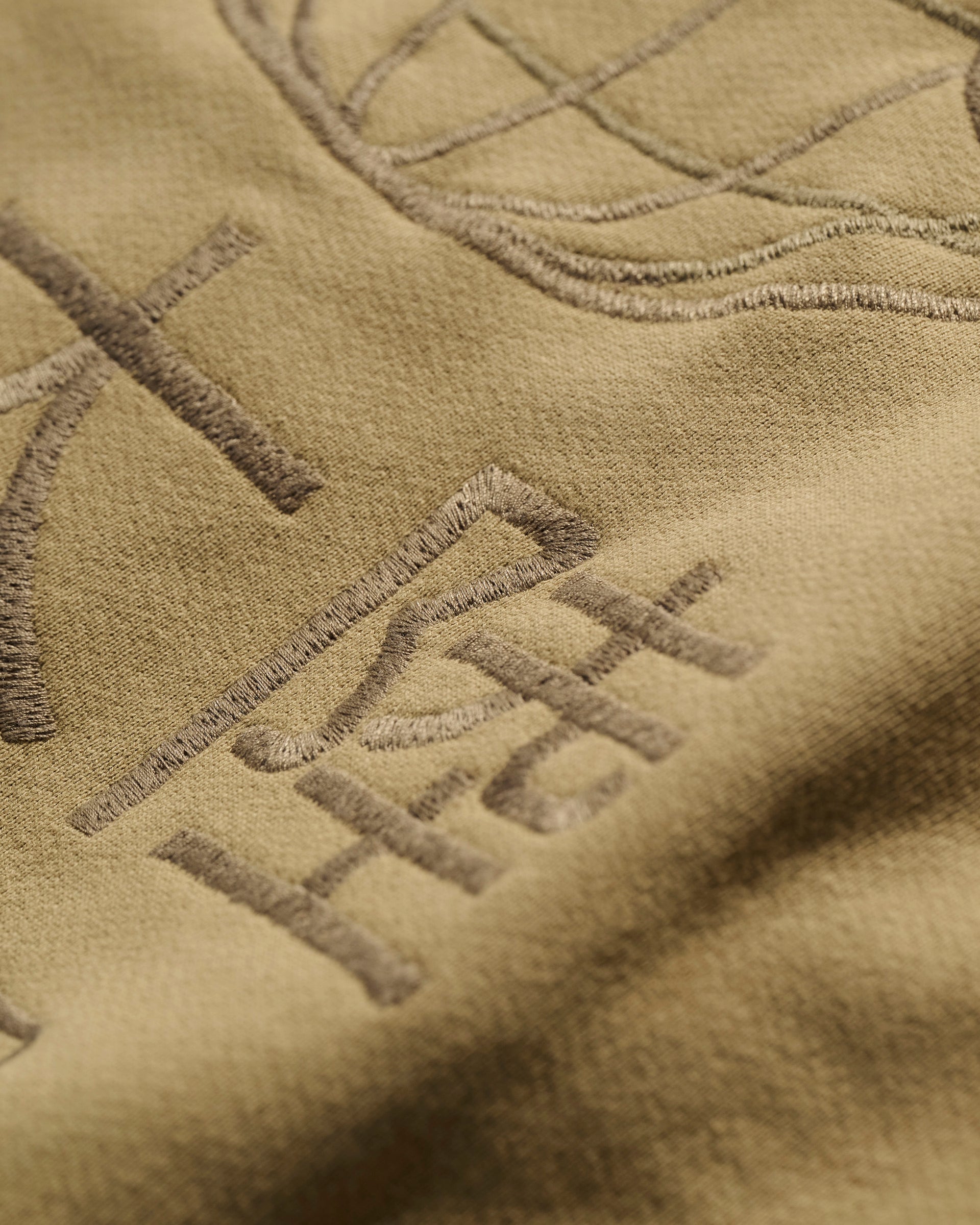 Close-up of The Camden Hoodie in Desert by T7C The Seven Continents, featuring tan fabric with raised, embroidered symbols, detailed stitching, and genuine leather accents.