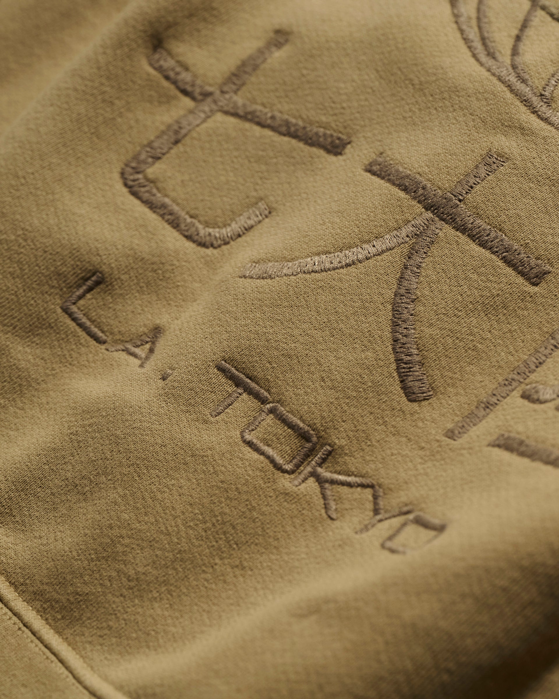Close-up of T7C The Seven Continents The Camden Hoodie in Desert: khaki fabric with embroidered Japanese characters, L.A. TOKYO text in matching thread, and genuine leather detailing for a refined finish.
