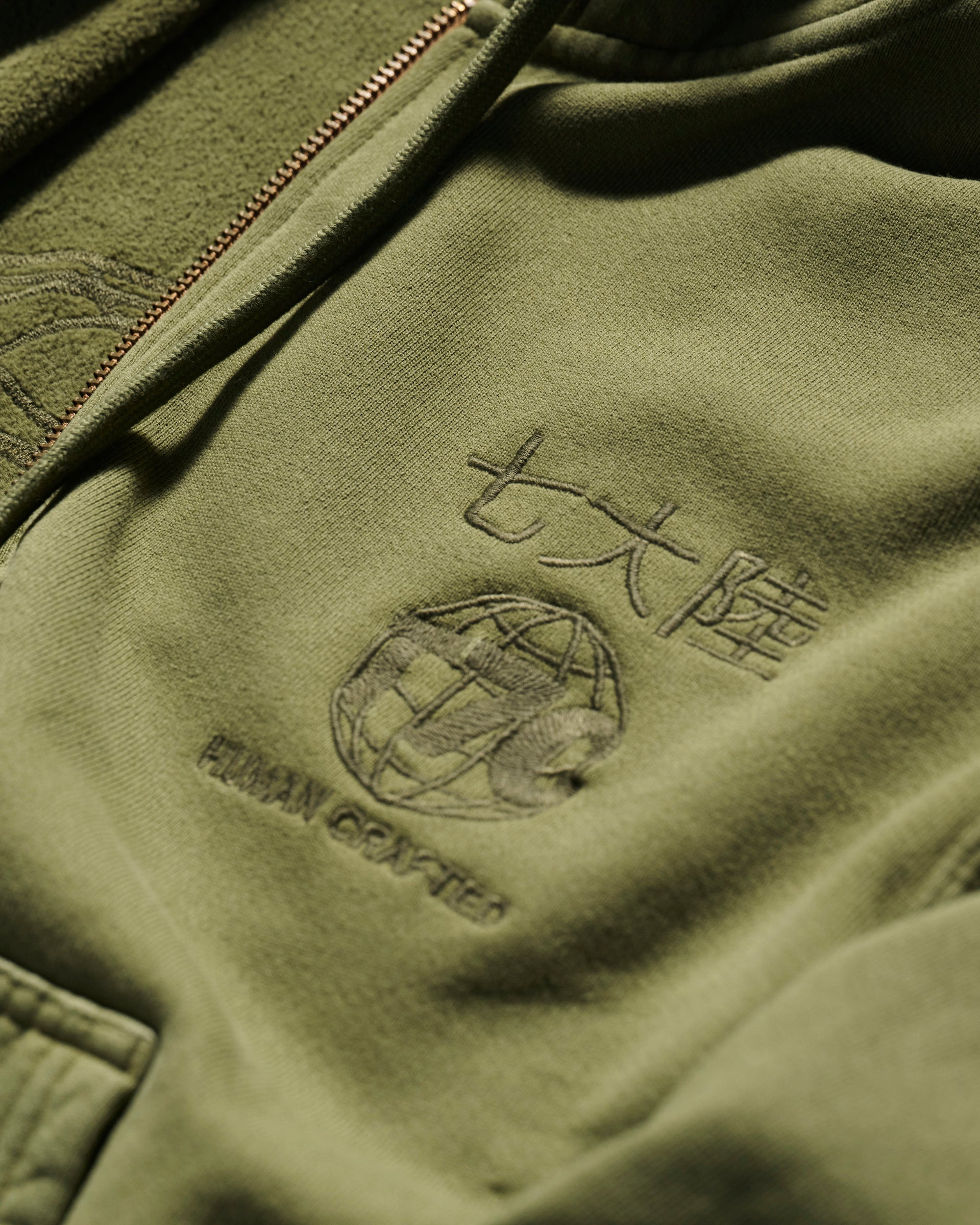 Close-up of The Camden Hoodie in Olive by T7C The Seven Continents, featuring embroidered HUMAN GRAFFITI text, a globe design, Asian characters, relaxed fit, front zipper, detailed stitching, and suede elbow patches.