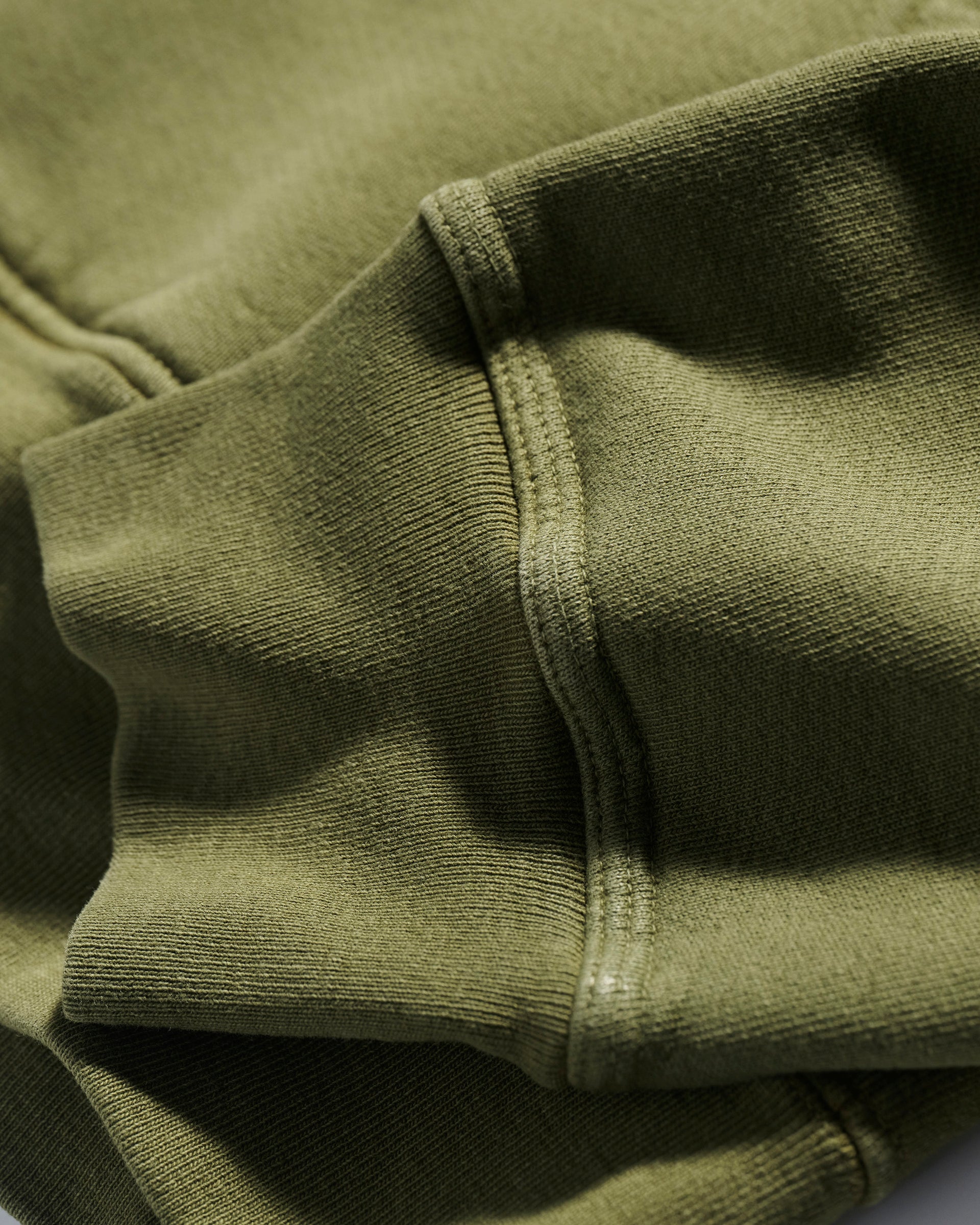 Close-up of olive green fabric, highlighting the textured surface and stitching detail of T7C The Seven Continents Camden Hoodie in Olive—a relaxed fit layer with soft folds and subtle style.