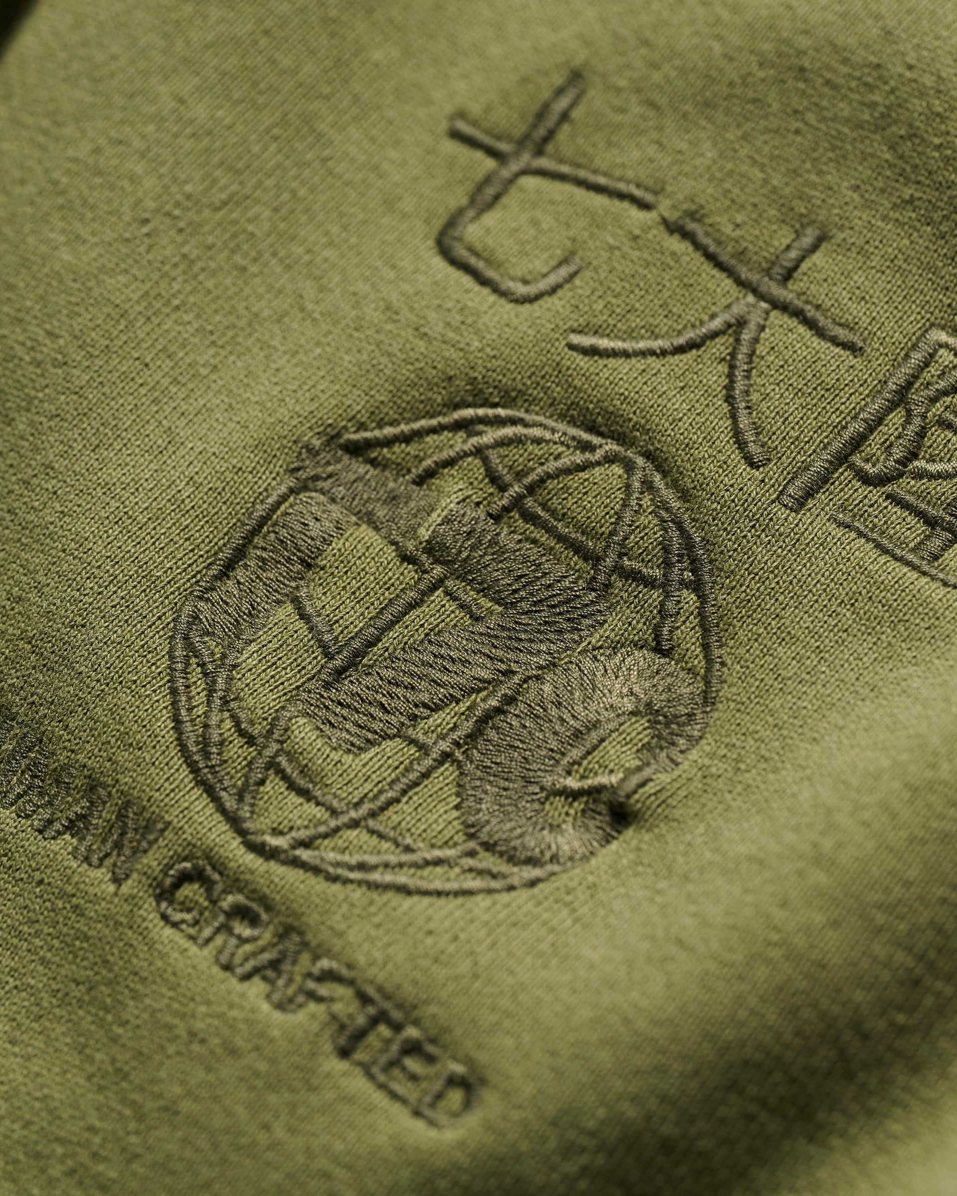 Close-up of olive green fabric from The Camden Hoodie in Olive by T7C The Seven Continents, featuring embroidered Asian characters, a globe motif, AN CRAFTED text, suede elbow patches, and detailed stitching on the textured material.