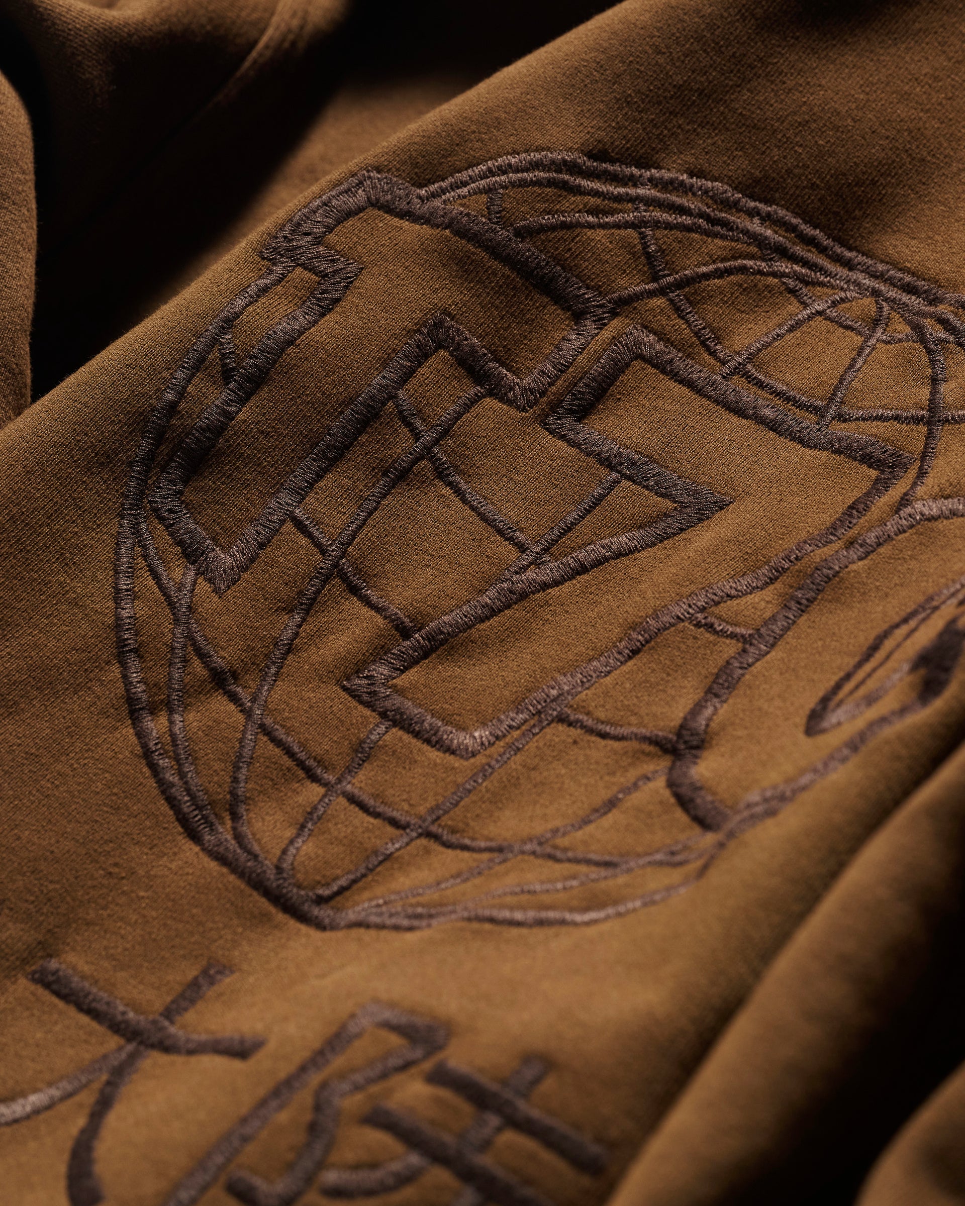 Close-up of The Camden Hoodie in Pecan by T7C The Seven Continents, showcasing brown fabric with an embroidered globe, geometric designs, stylized characters, and suede elbow patches for a relaxed, detailed look.