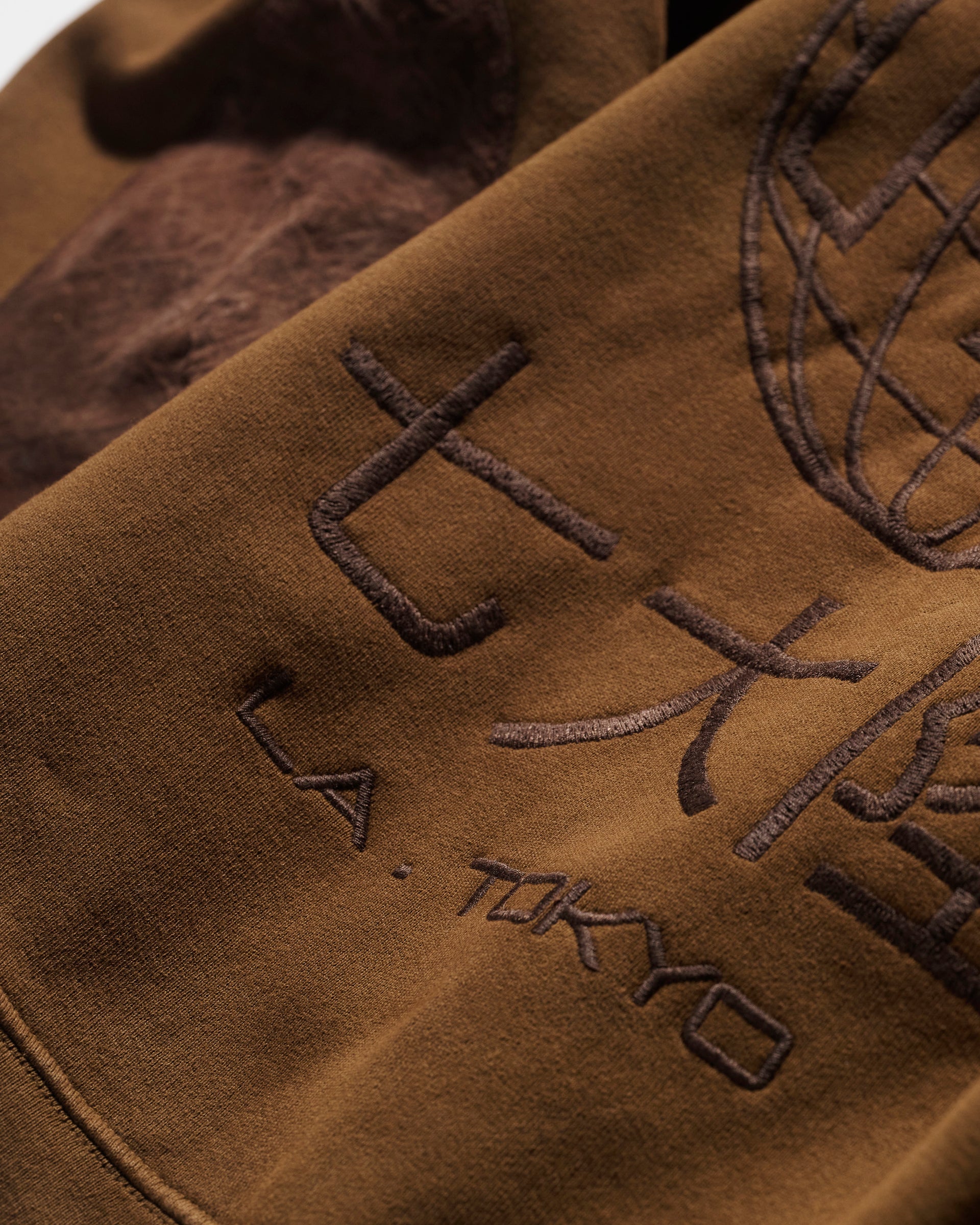 A close-up of The Camden Hoodie in Pecan by T7C The Seven Continents shows brown fabric with L.A. TOKYO and large Japanese characters embroidered beside a partial stitched globe, all in a darker brown thread for subtle detail.