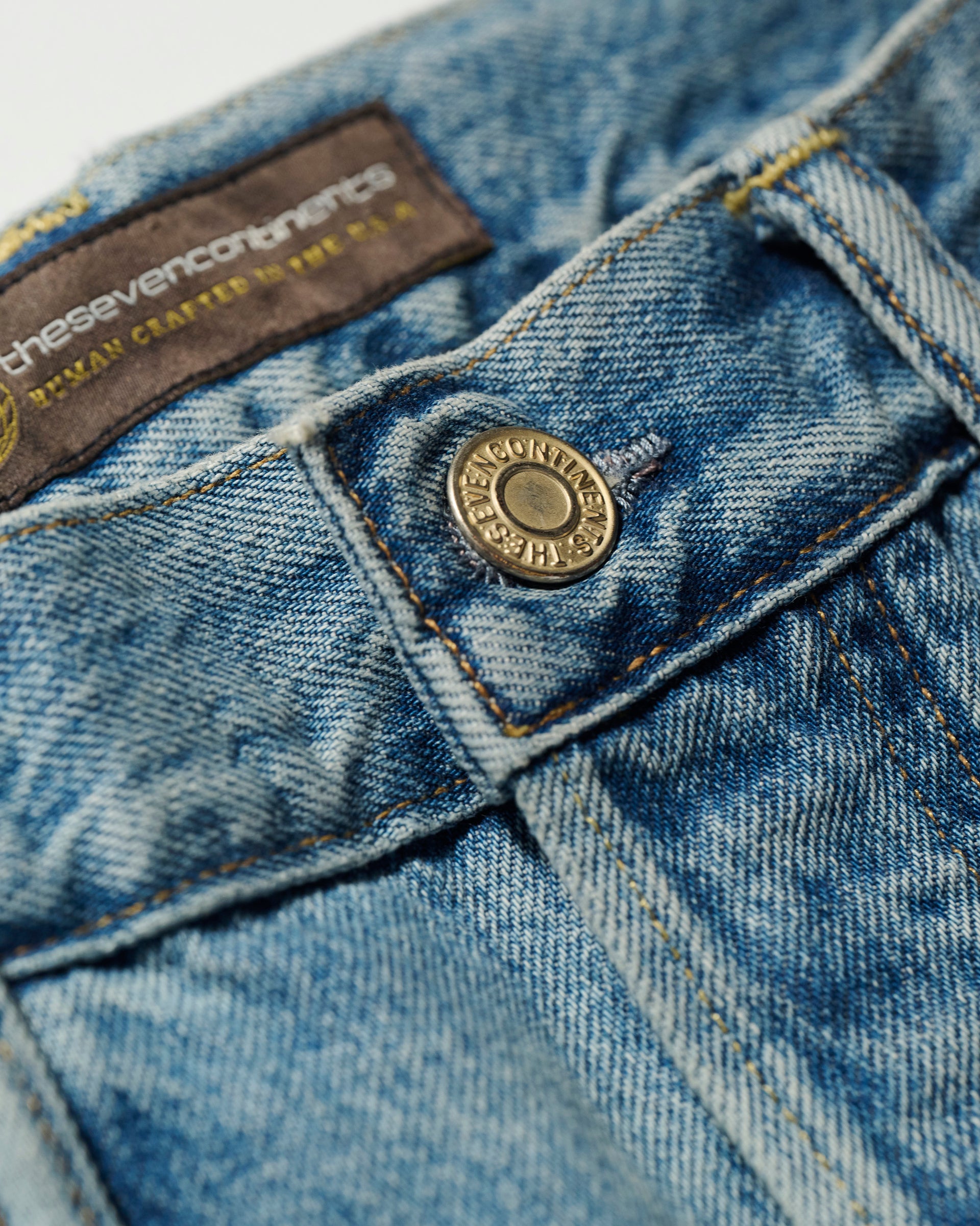 Close-up of the waistband on The Charlie Jean in Zuma by T7C The Seven Continents, featuring a metal button, belt loops, and a brown inside label with yellow text. The blue selvedge denim shows a faded, textured look.