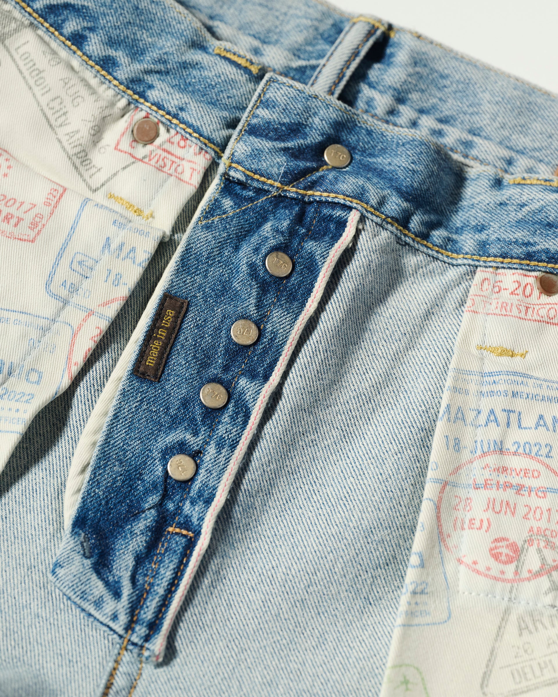 Close-up of T7C The Seven Continents The Charlie Jean in Zuma: blue Japanese selvedge denim with exposed metal button fly and a vibrant travel stamp pattern lining, displaying destinations and dates.