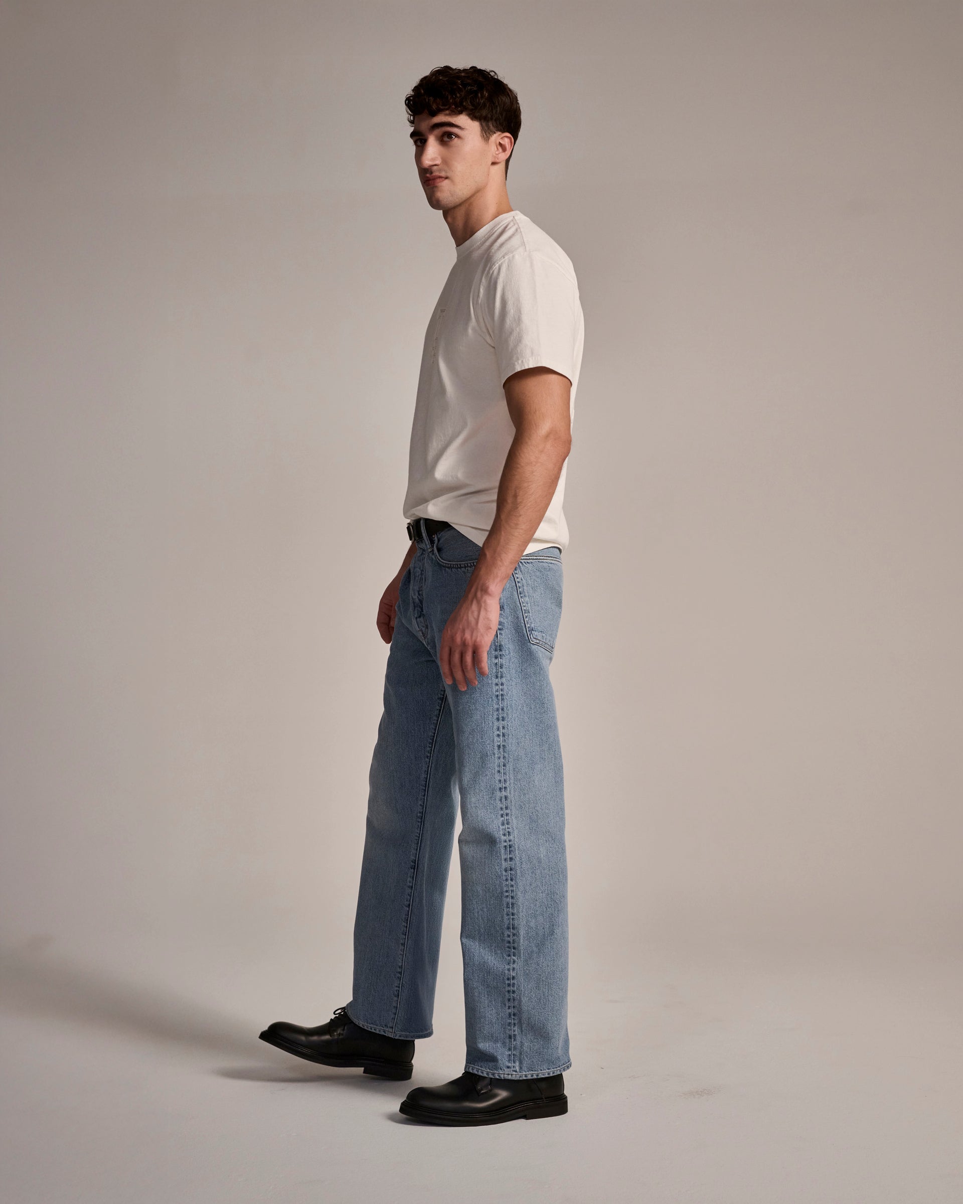 A man with short dark hair stands sideways against a plain background, wearing a white T-shirt, black shoes, and T7C’s The Cinch Jean in Mykonos.