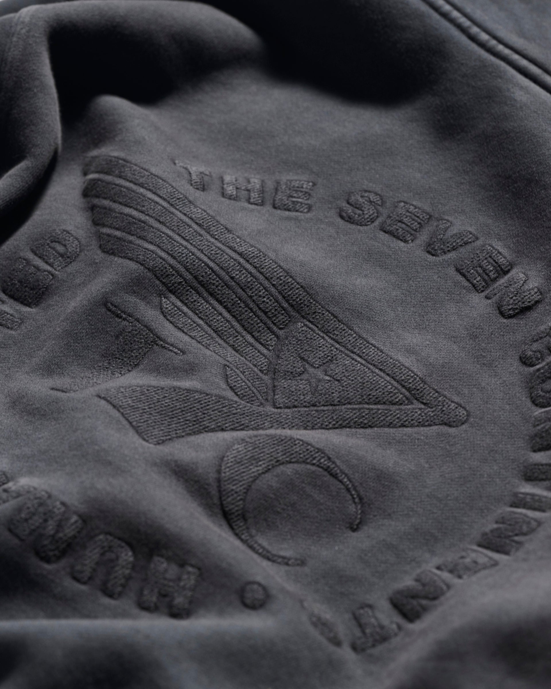 Close-up of THE DAX IN VINTAGE BLACK by T7C: dark fabric with embossed embroidery of an eye, lines above, a crescent moon below, and partially visible circular text.