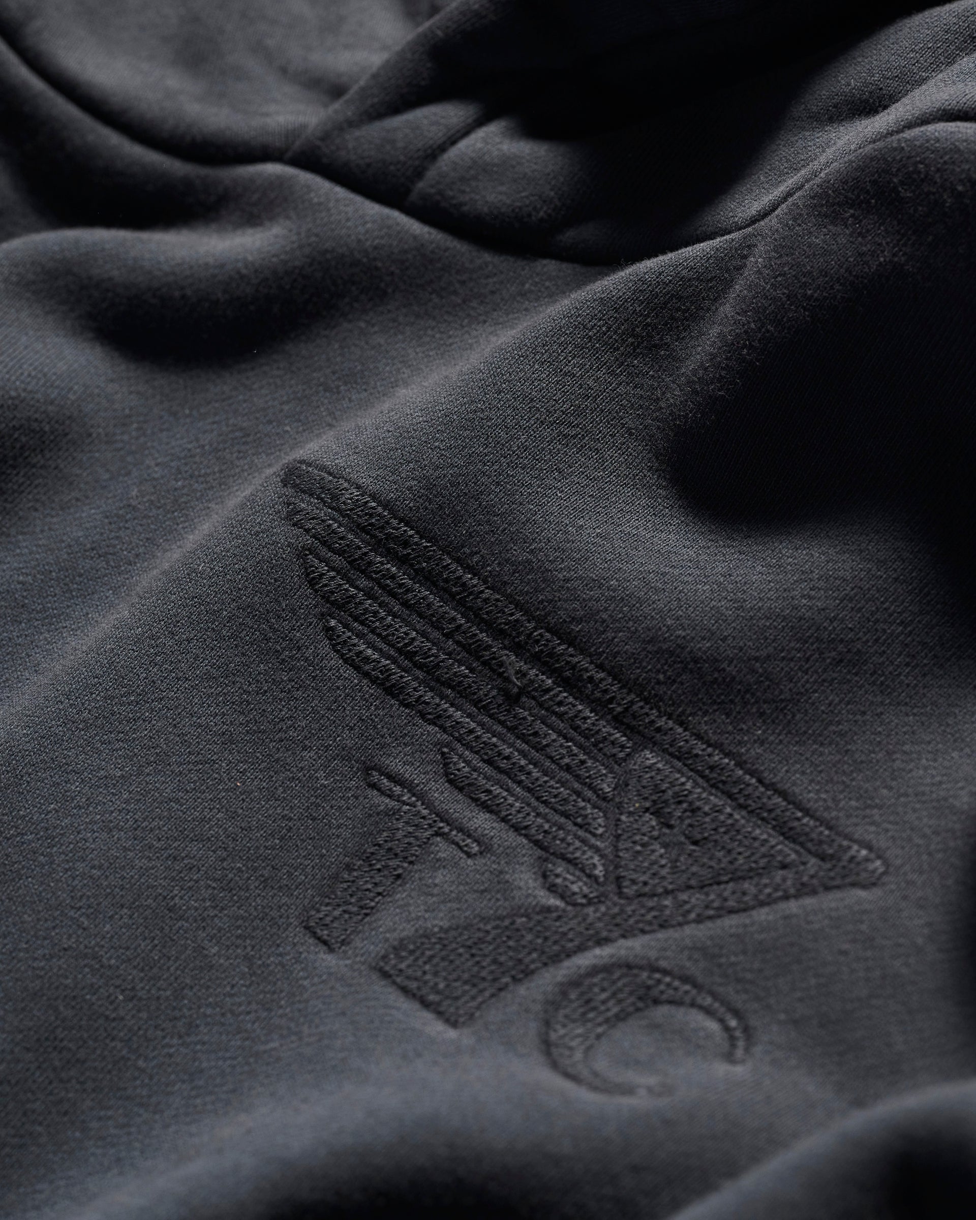 Close-up of THE DAX IN VINTAGE BLACK hoodie by T7C, featuring a dark fabric with an embossed minimalist wing graphic and the letters T C.