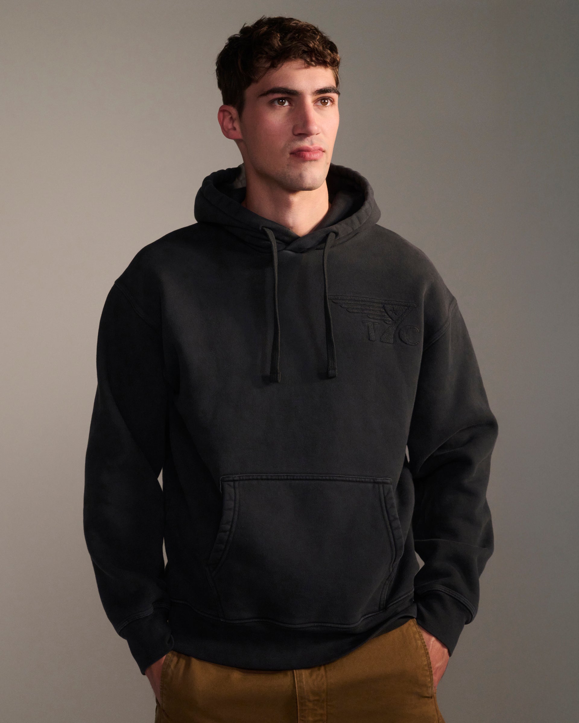 A young man wears THE DAX IN VINTAGE BLACK hoodie by T7C with tan pants, standing against a plain background and looking slightly to the side with a neutral expression.