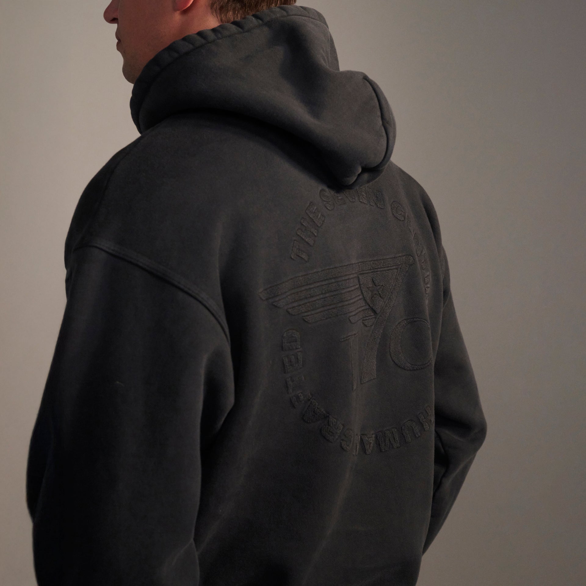 A person wearing THE DAX IN VINTAGE BLACK hoodie by T7C, featuring a raised winged logo and the number 10 on the back, stands slightly turned away from the camera against a neutral background.