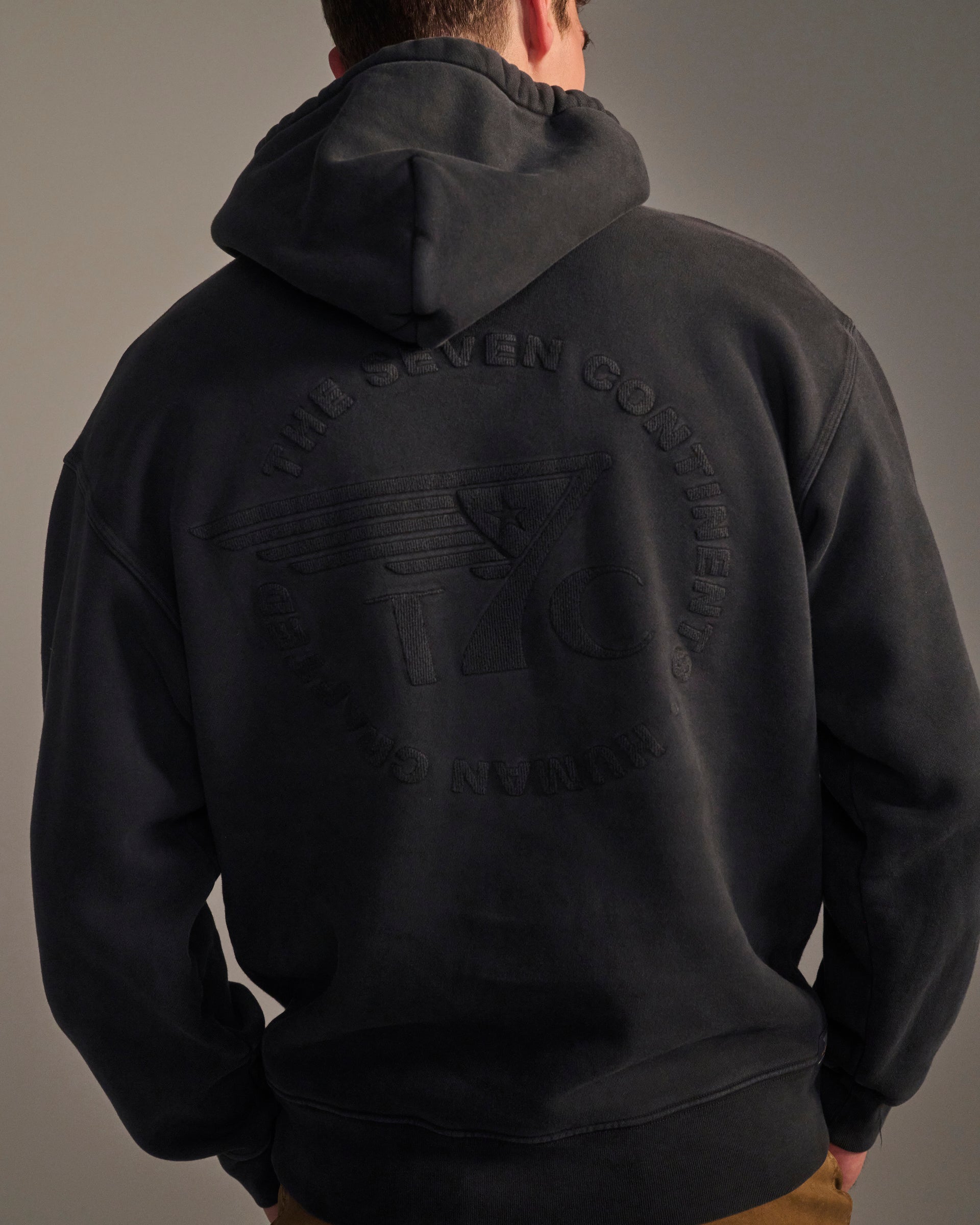 A person faces away from the camera, wearing T7C's THE DAX IN VINTAGE BLACK hoodie featuring THE SEVEN CONTINENTS text and a circular logo on the back.
