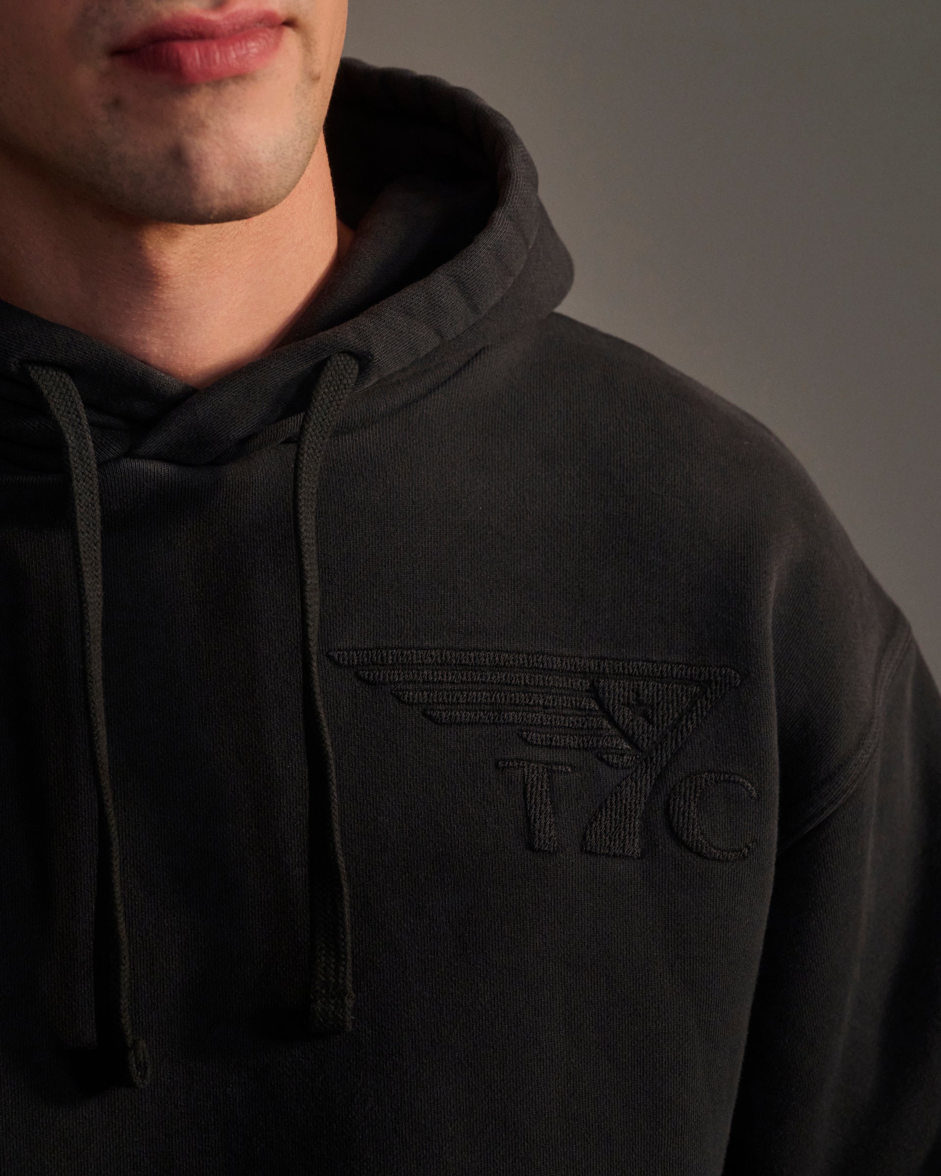 A person wearing T7C's THE DAX IN VINTAGE BLACK hoodie, showcasing a raised embroidered YC logo with stylized wings on the left chest. Only the lower half of their face and torso are visible.
