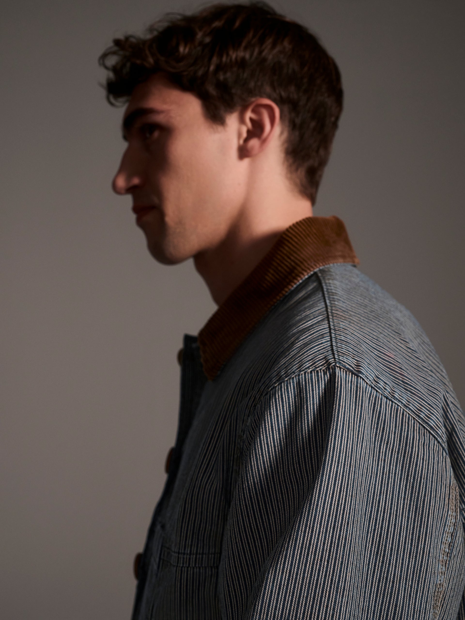 A detail image of a male model wearing a blue and white railroad stripe jacket with a corduroy collar