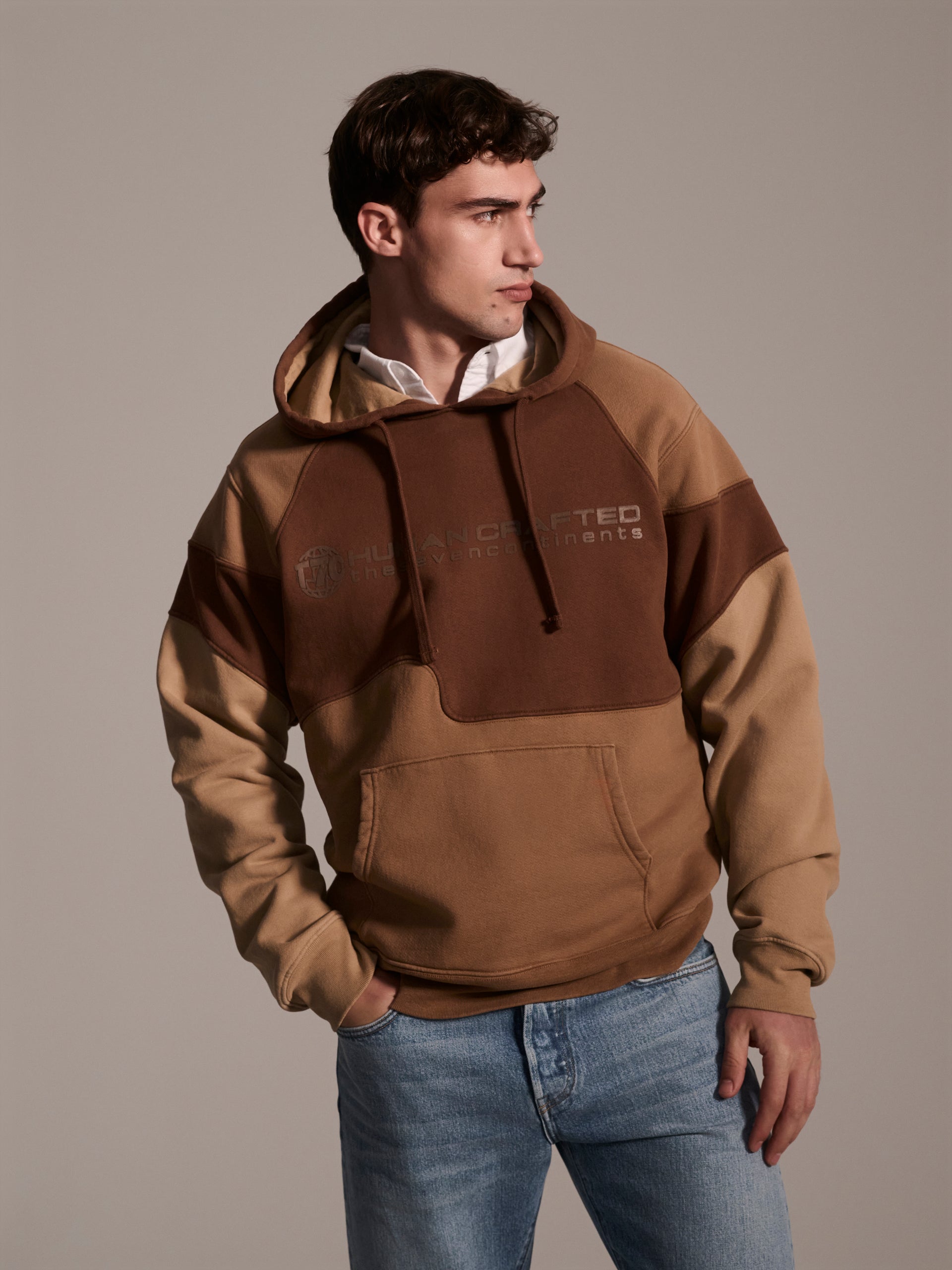 A front image of a male model wearing a color blocked pull-over hoodie in brown tones featuring a printed logo 