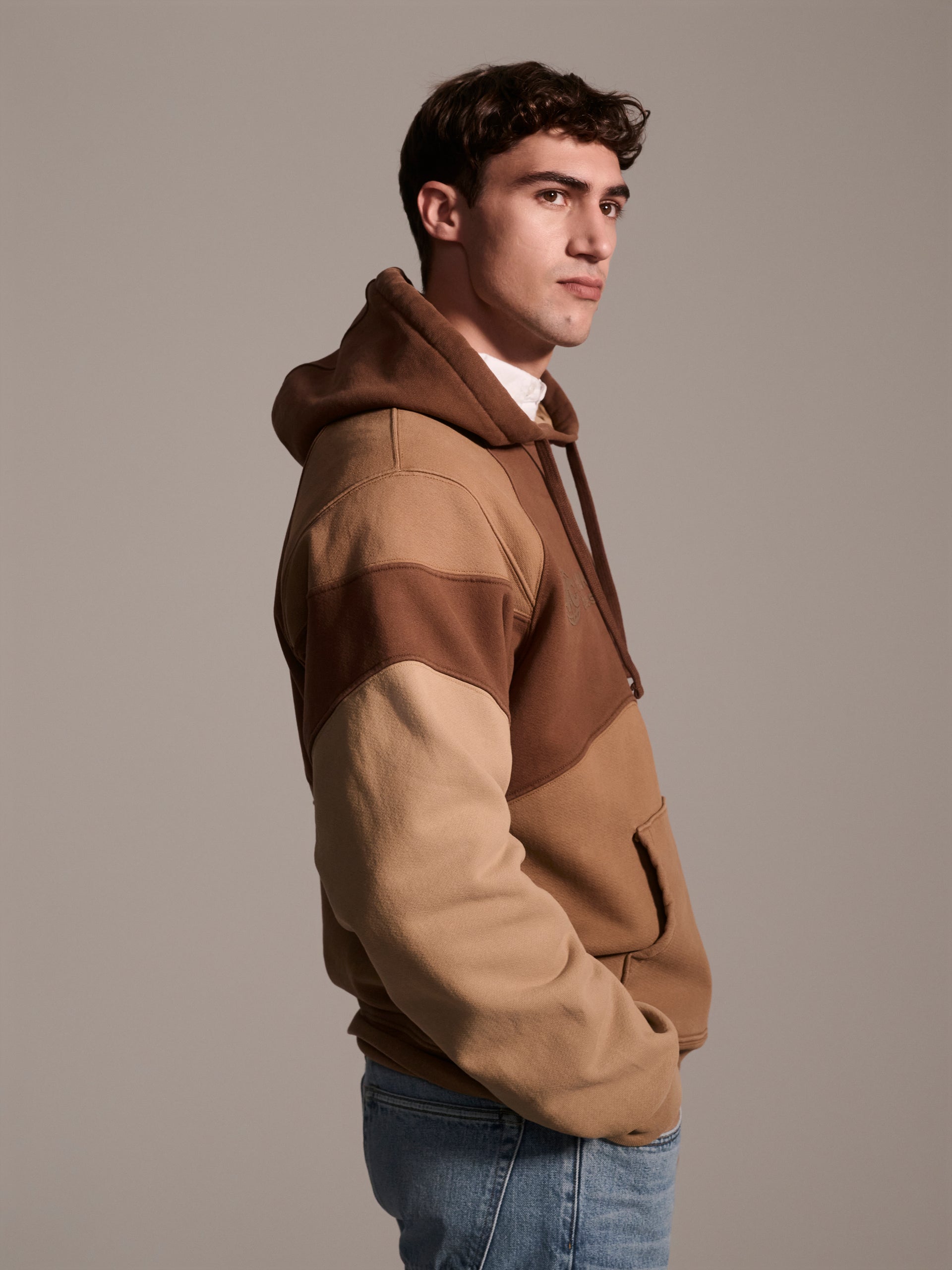 A profile image of a male model wearing a color blocked pull-over hoodie in brown tones featuring a printed logo 