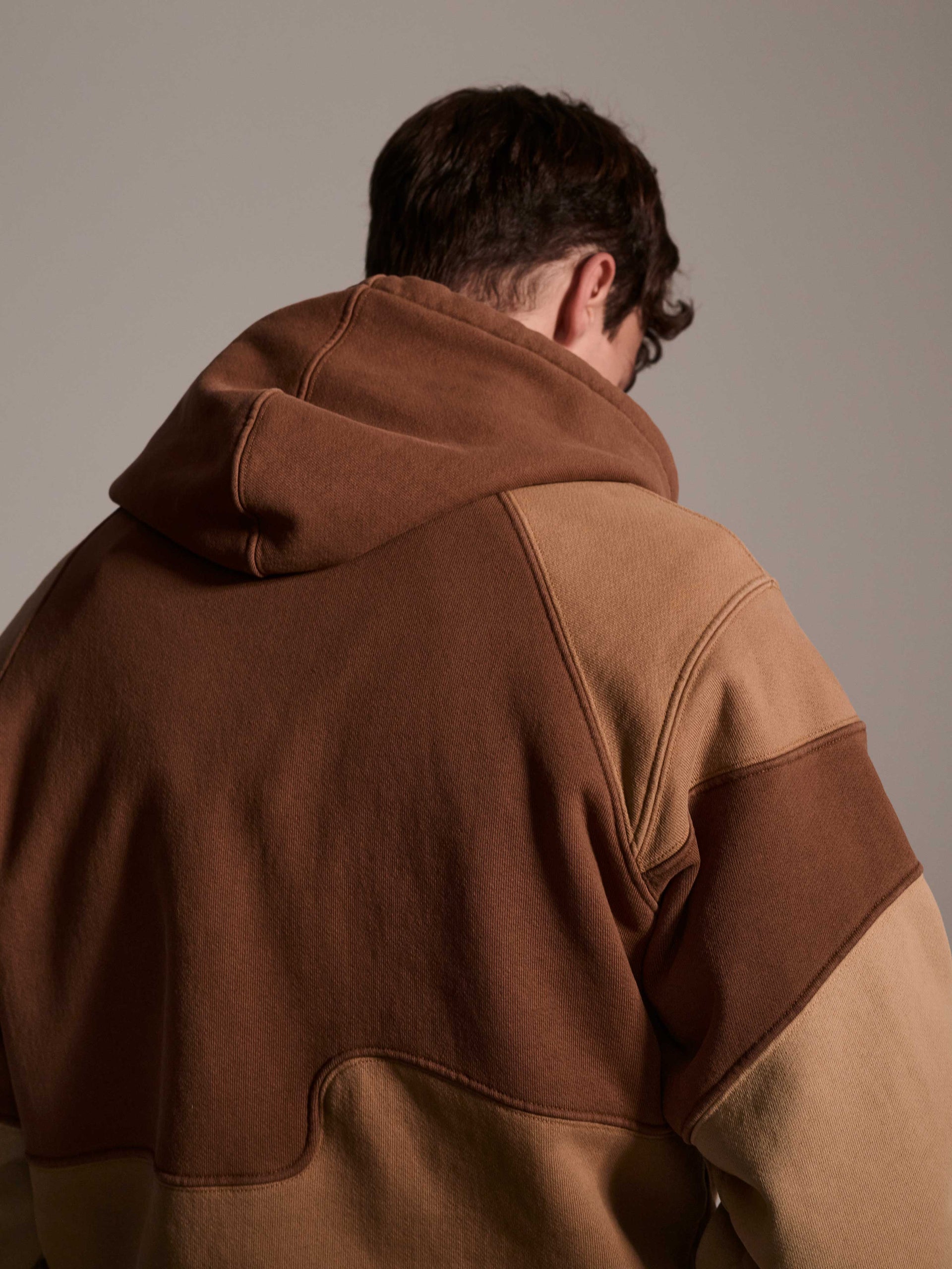 A back detail image of a male model wearing a color blocked pull-over hoodie in brown tones featuring a printed logo 