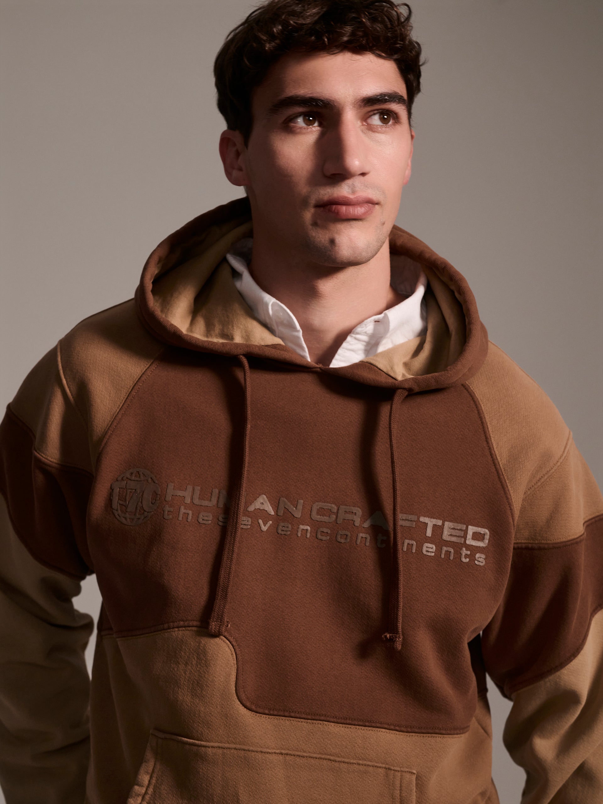 A detail image of a male model wearing a color blocked pull-over hoodie in brown tones featuring a printed logo 