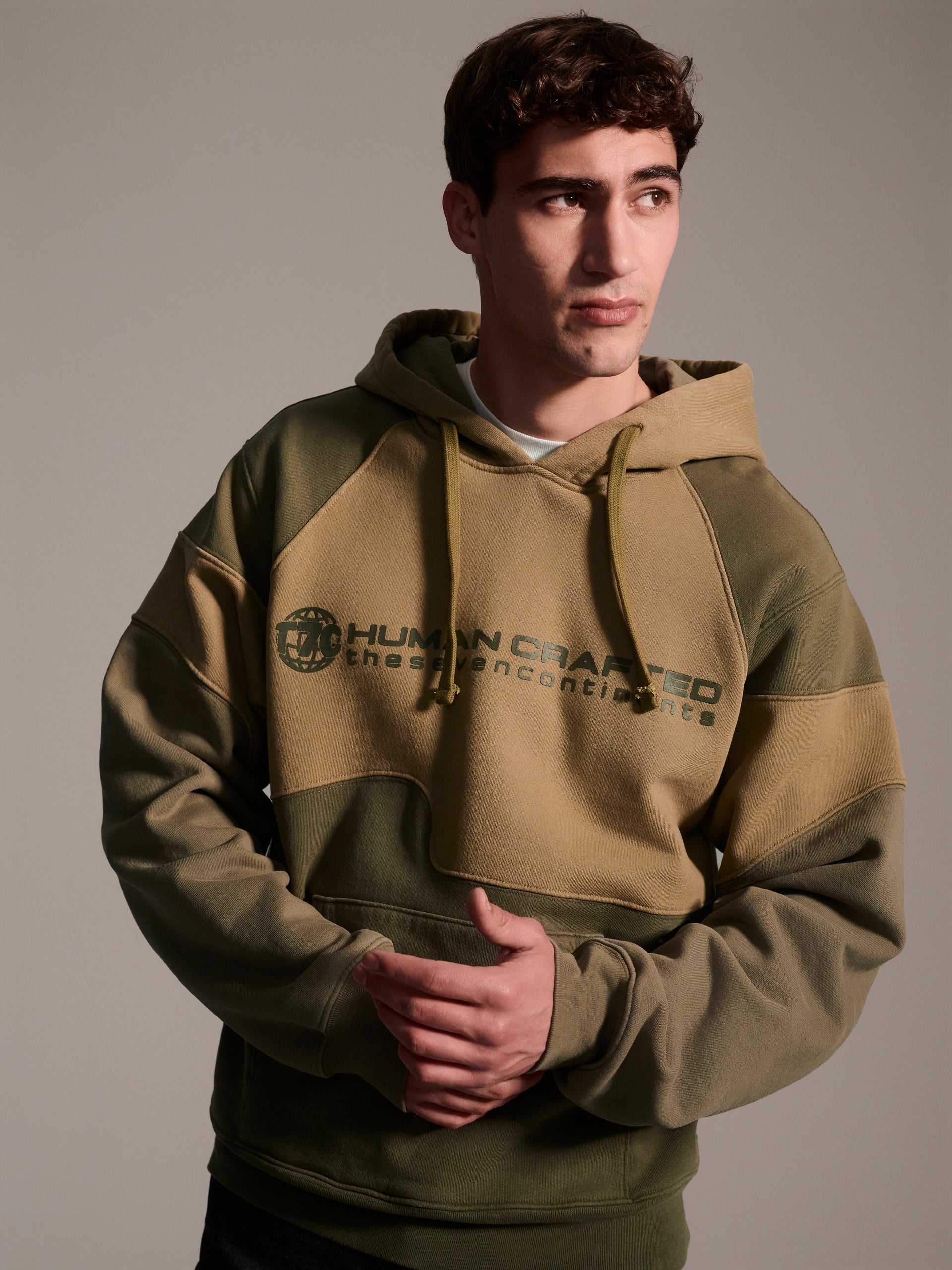 A front image of a male model wearing a color blocked pull-over hoodie in green tones featuring a printed logo 