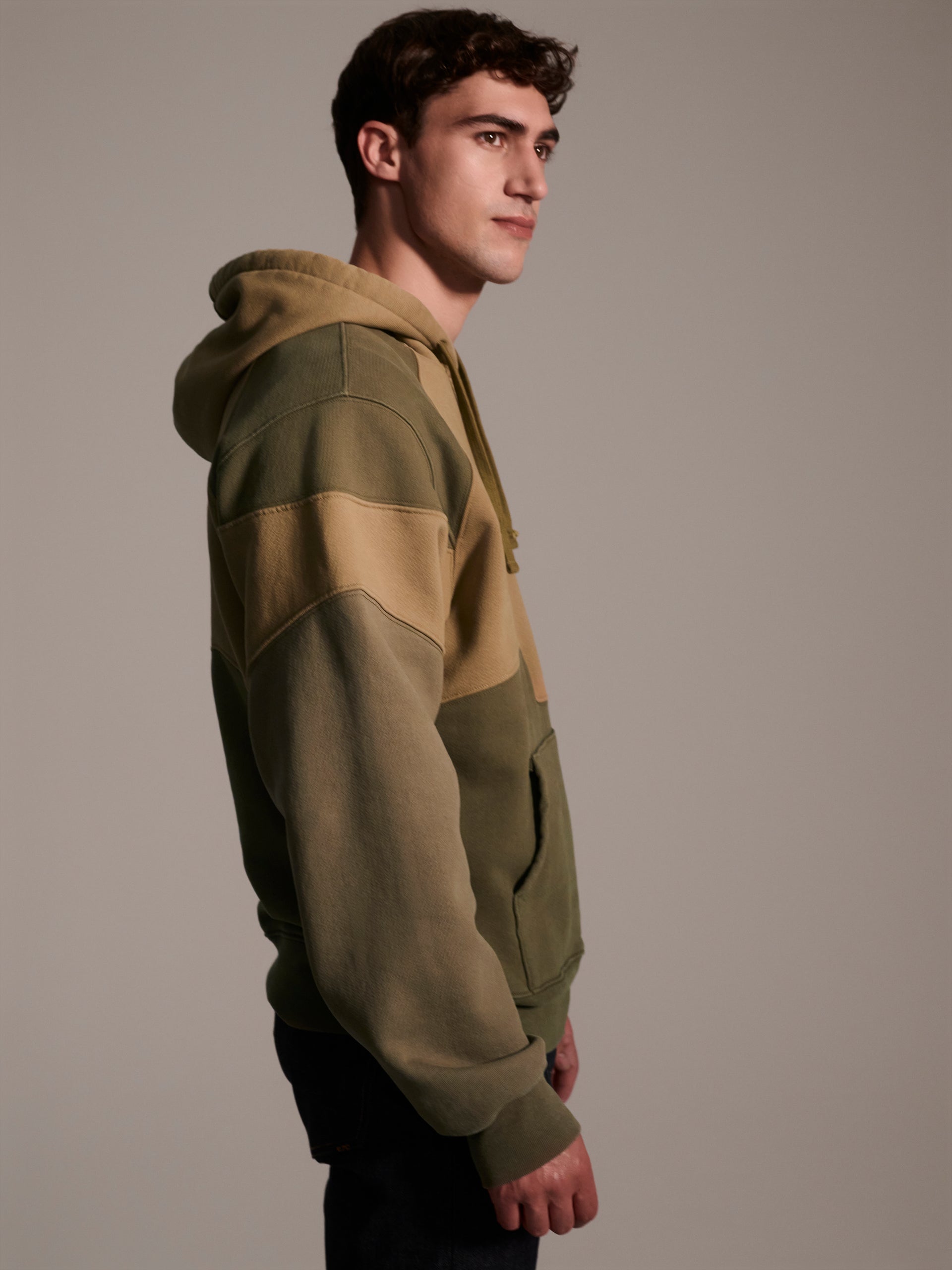 A profile image of a male model wearing a color blocked pull-over hoodie in green tones featuring a printed logo 