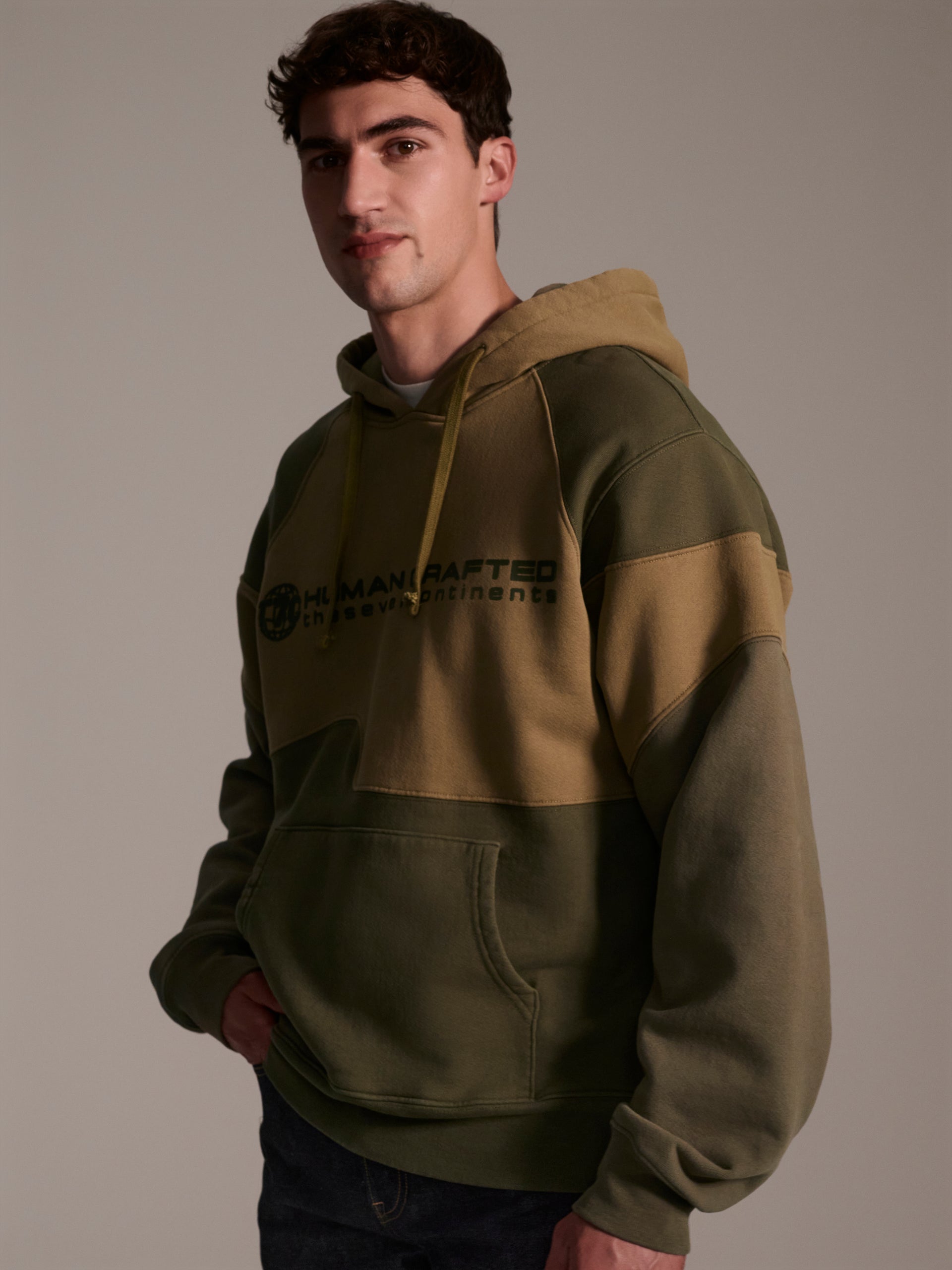 A profile image of a male model wearing a color blocked pull-over hoodie in green tones featuring a printed logo 