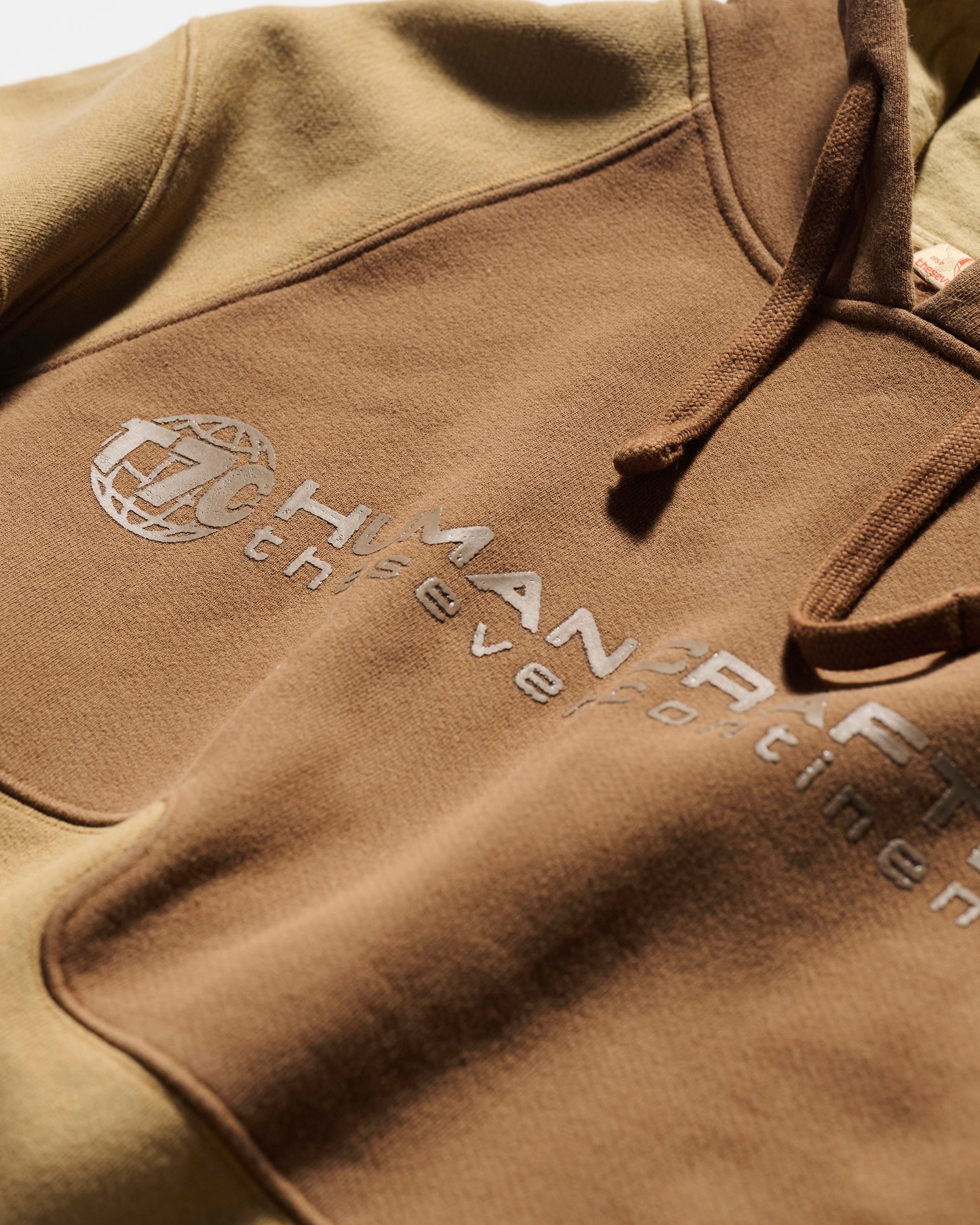 A close-up of T7C The Seven Continents Hunter Hoodie in Terra, featuring HUMAN RACE and a globe embroidered on the chest, with gradient tan-to-brown fabric, visible drawstrings, and camo-inspired seam detail.
