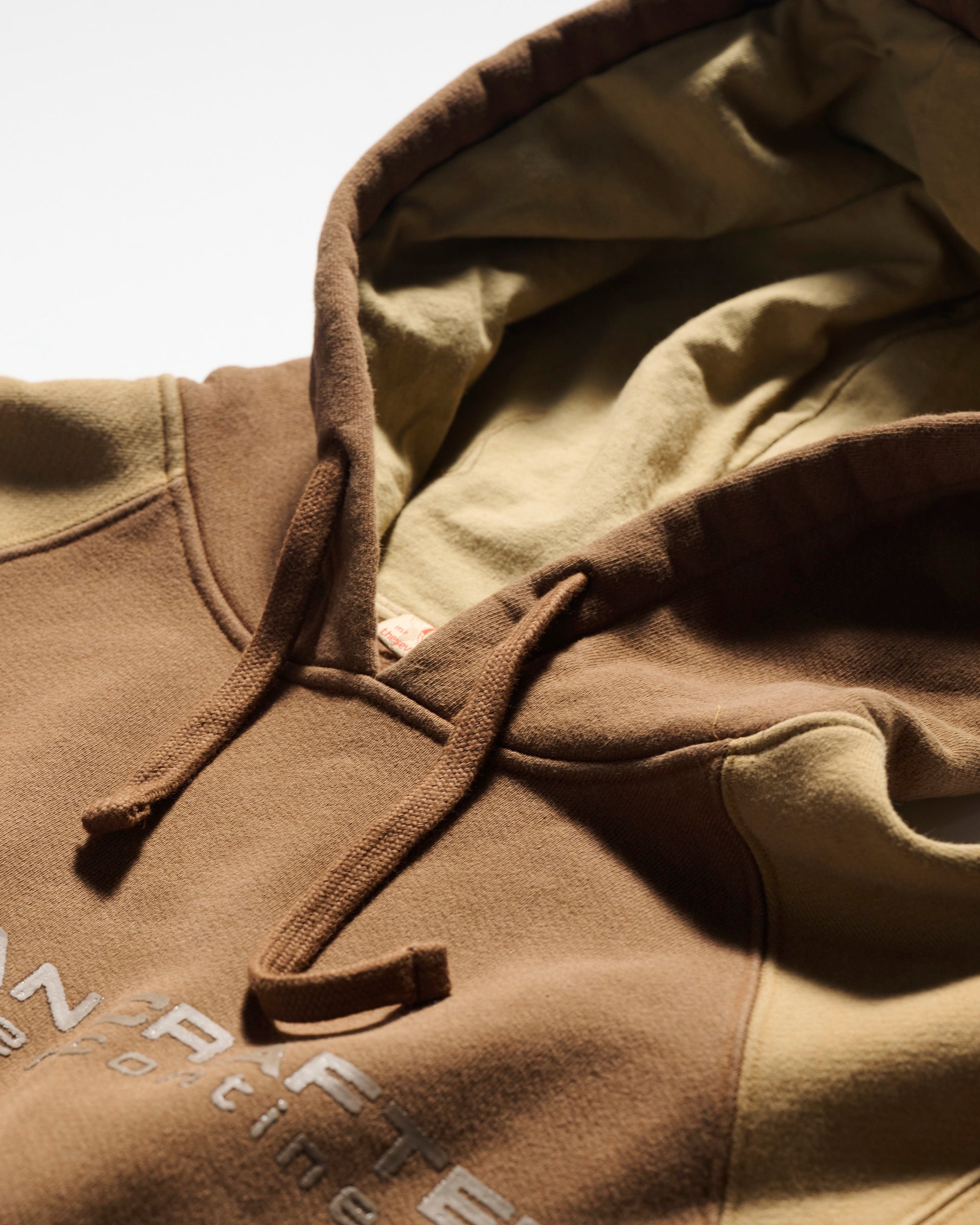 Close-up of T7C The Seven Continents Hunter Hoodie in Terra, a brown and beige pullover with drawstrings, partial embroidered text, camo-inspired seam, and soft fabric laid flat on a light surface.
