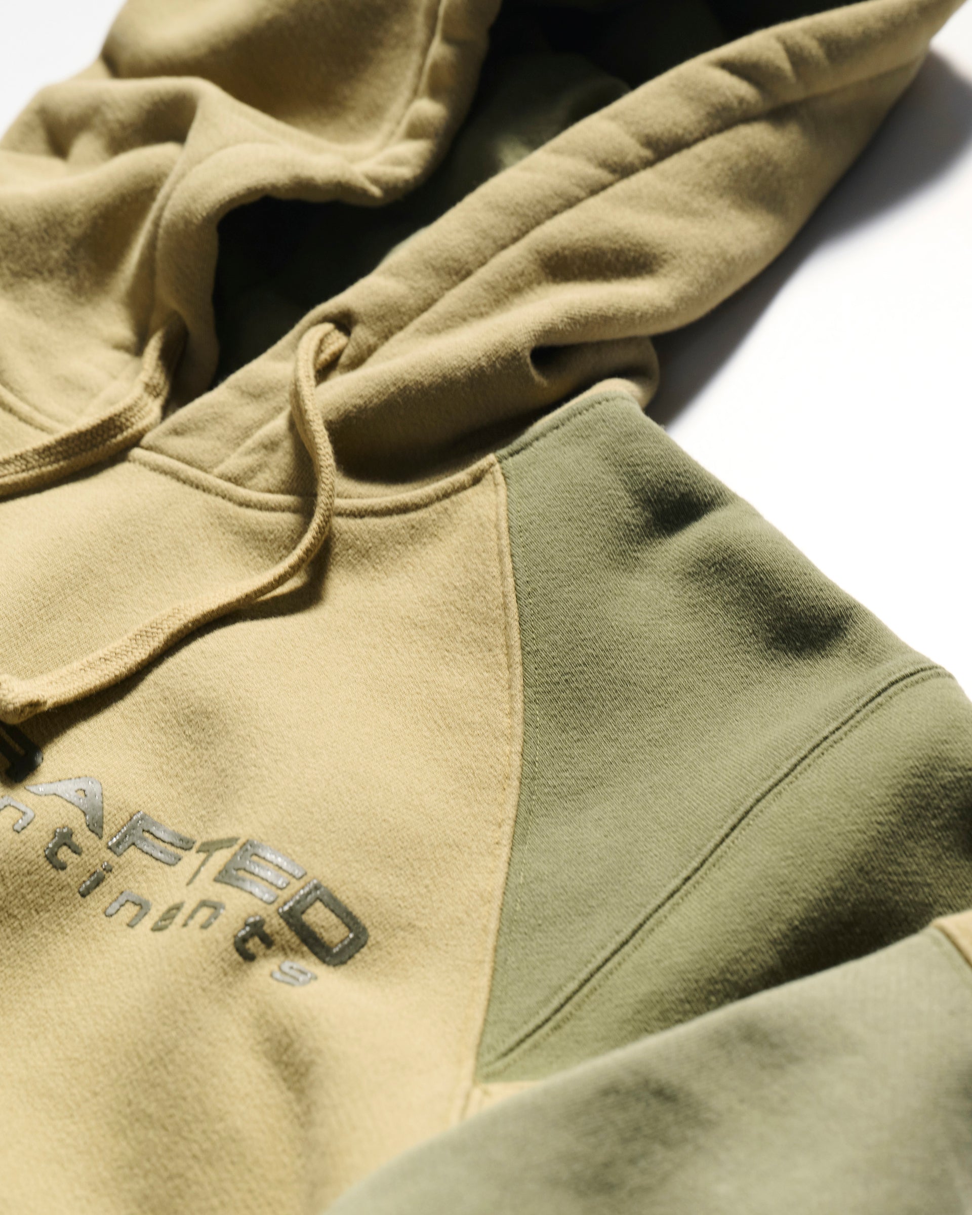 A close-up of The Hunter Hoodie in Verde by T7C The Seven Continents, featuring beige and olive green tones, drawstrings, camo-inspired seams, and a shiny printed chest logo, shown against a white background.