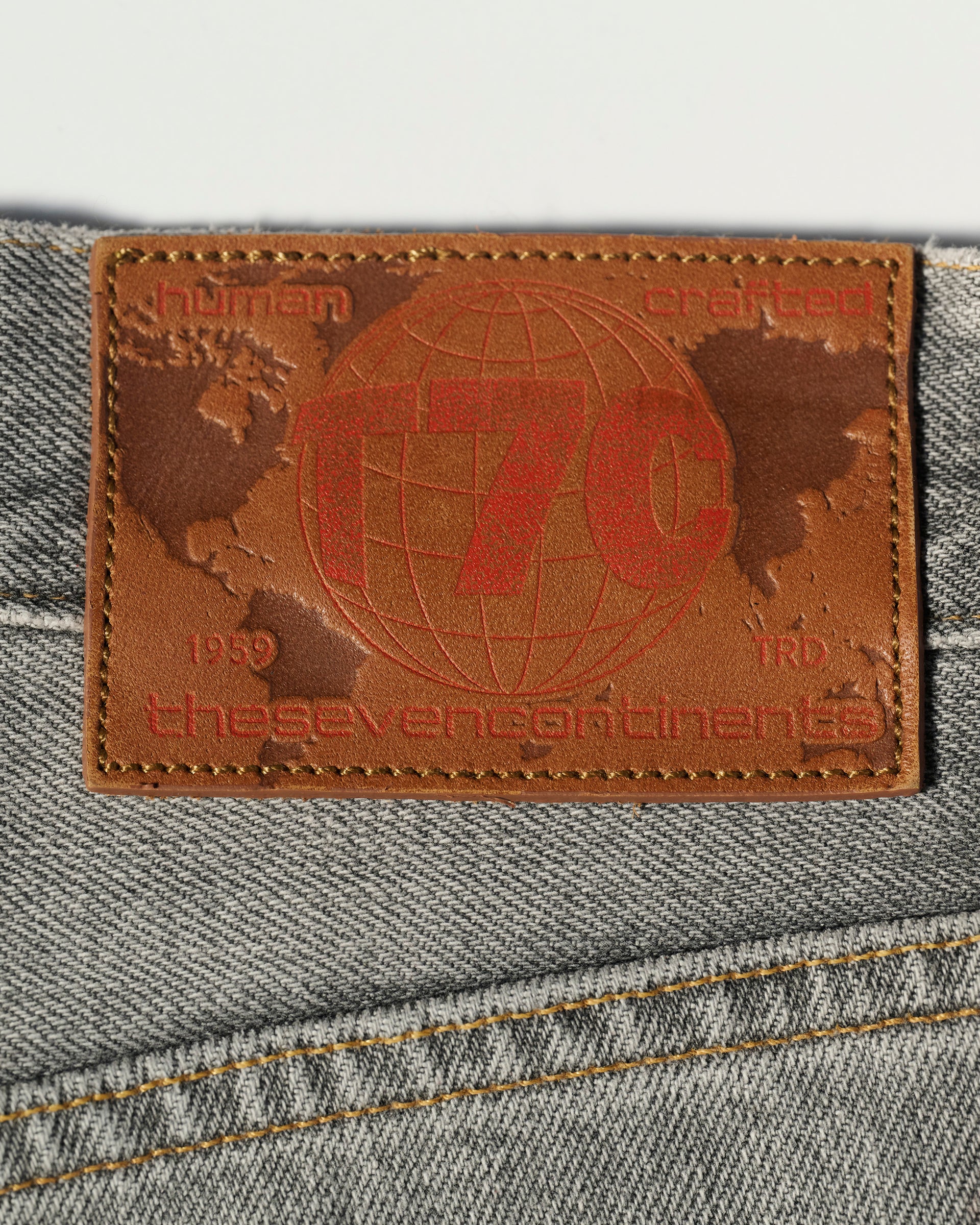 The Jack Jean in Cloud by T7C features a brown leather patch on gray organic Turkish cotton denim with a red globe, the text T7C, human crafted, the seven continents, and the years 1959 and TRD.