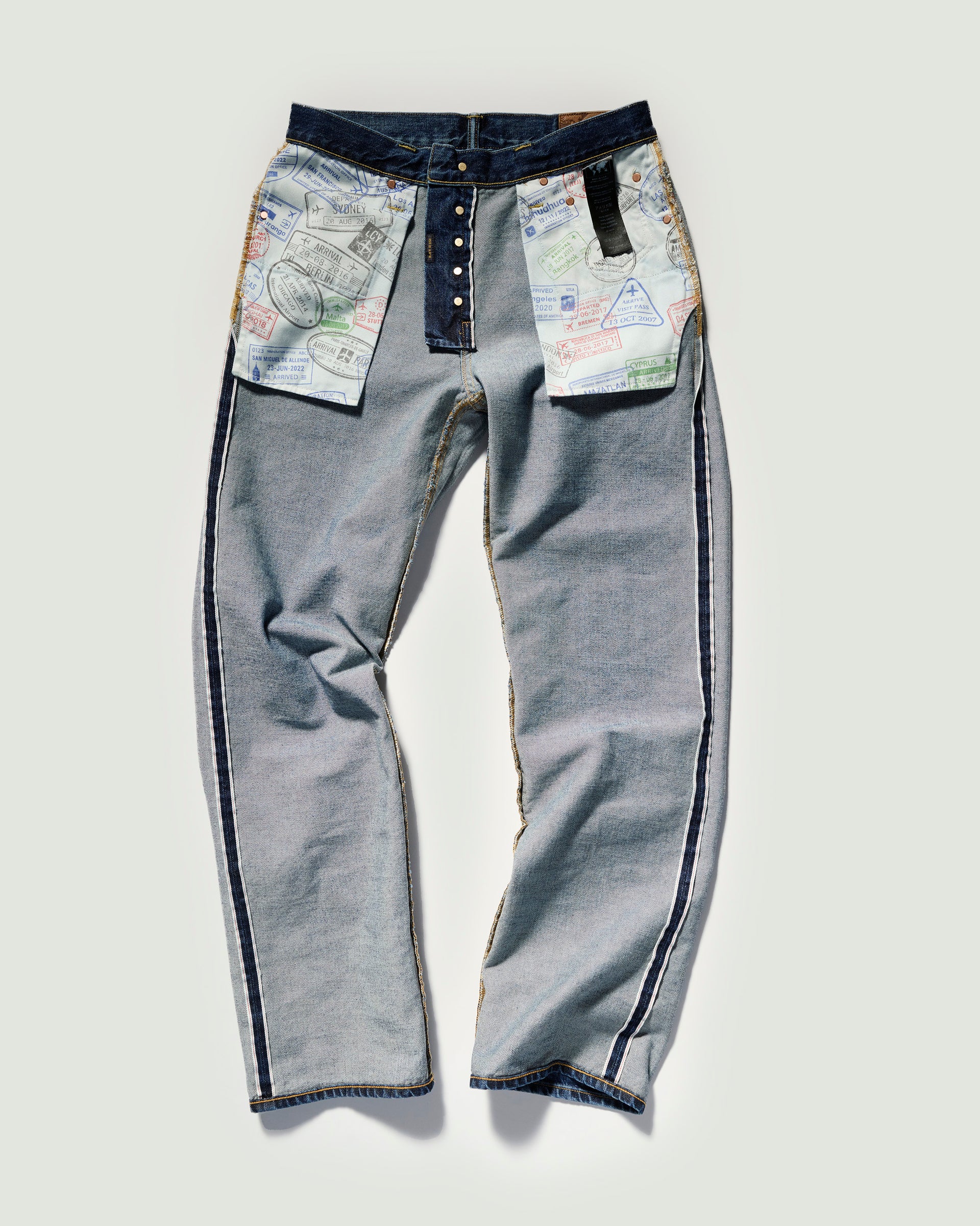 The Jack Jean in Tokyo Medium by T7C features light blue denim turned inside out, showcasing passport stamp prints on pocket linings and button fly, plus dark denim trim on the waistband and outer seams for a vintage feel.