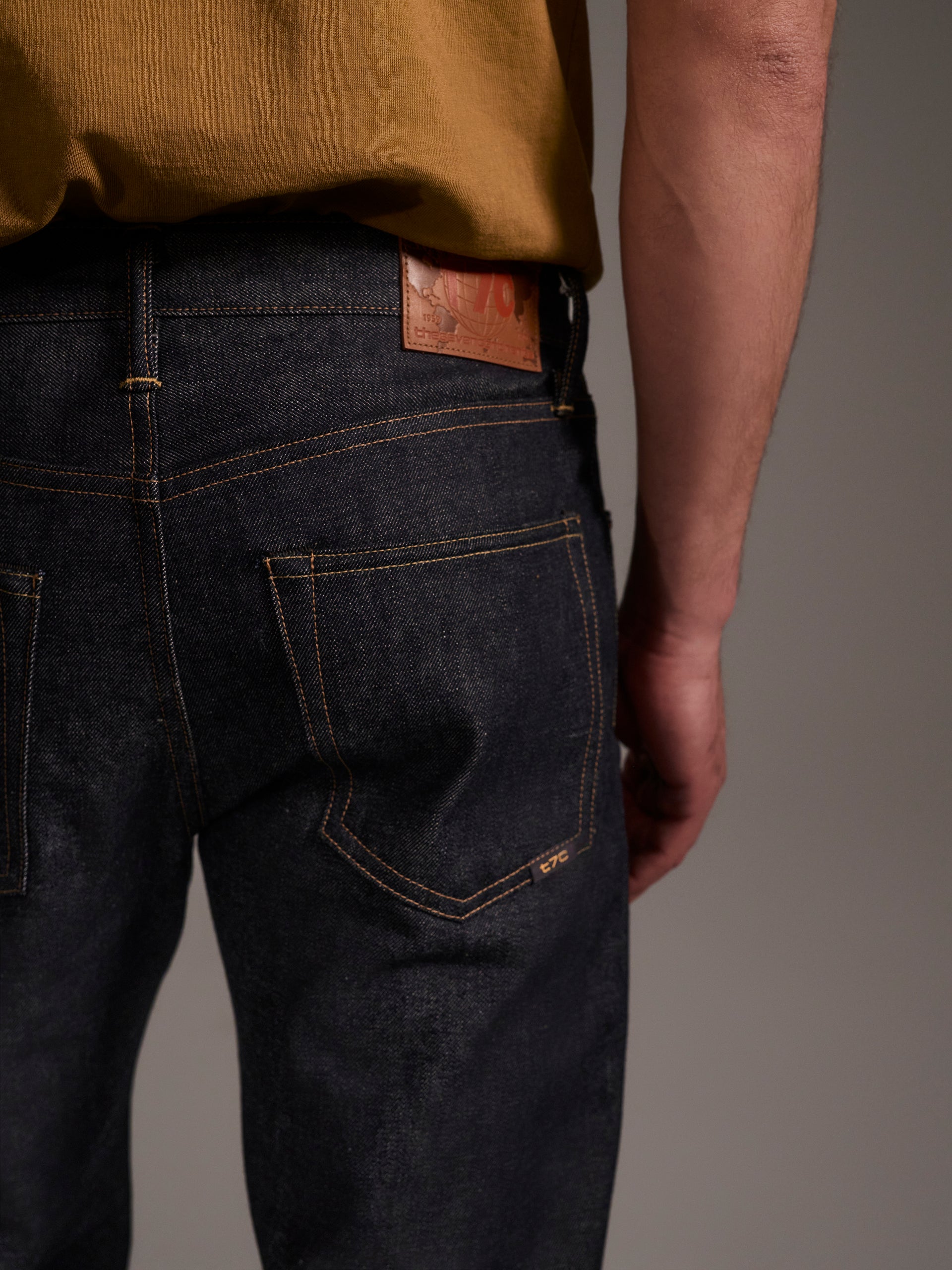 A back detail image of a male model wearing dark blue relaxed fit raw denim jeans with contrast stitching