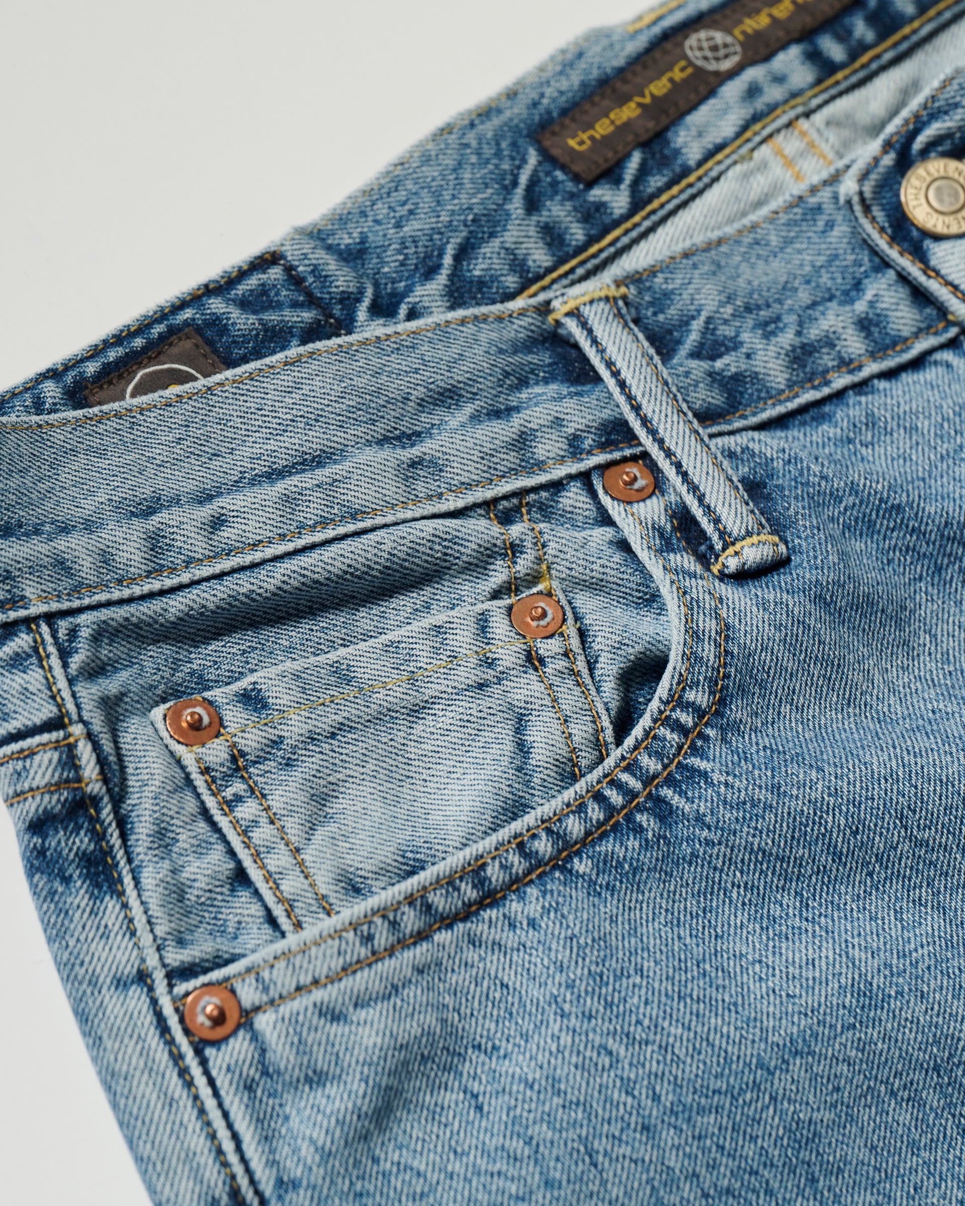 Close-up of the front pocket on The Jack Jean in Zuma by T7C The Seven Continents, highlighting the coin pocket, copper rivets, belt loops, stitching, part of the waistband with visible brand label, and vintage-textured Japanese denim.